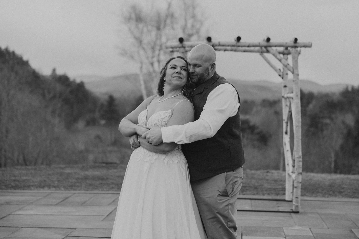Black and white photo of a couple embracing during an outdoor wedding ceremony with mountain backdrop.