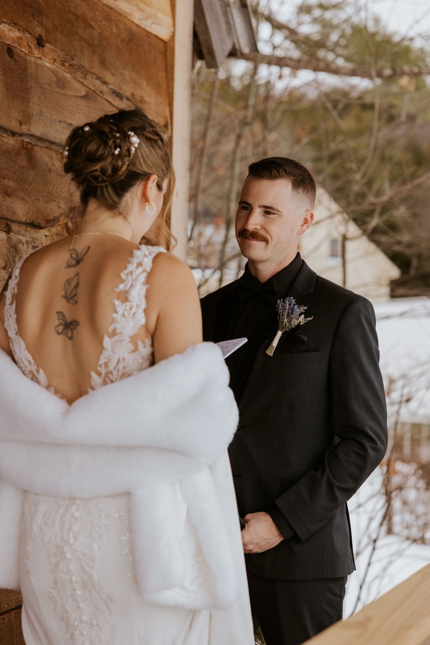 Groom in black tuxedo with mustache smiles at bride in lace backless wedding dress with fur wrap outdoors in winter.