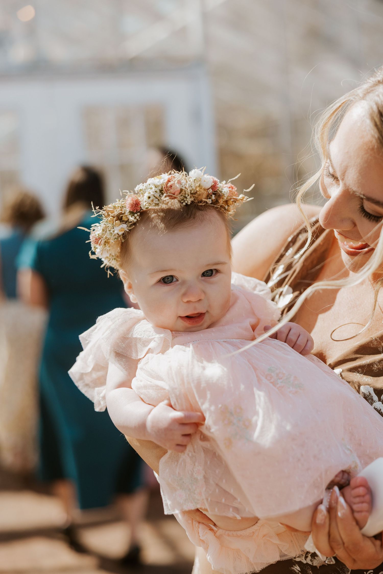 A baby wearing a pink dress and floral crown smiles during a special celebration.