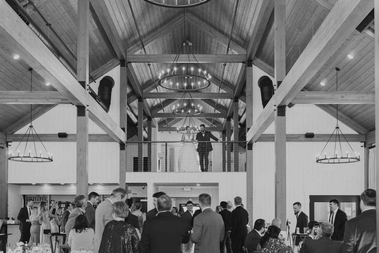 Black and white photo of rustic barn wedding reception with wooden beam ceiling and chandelier lighting.