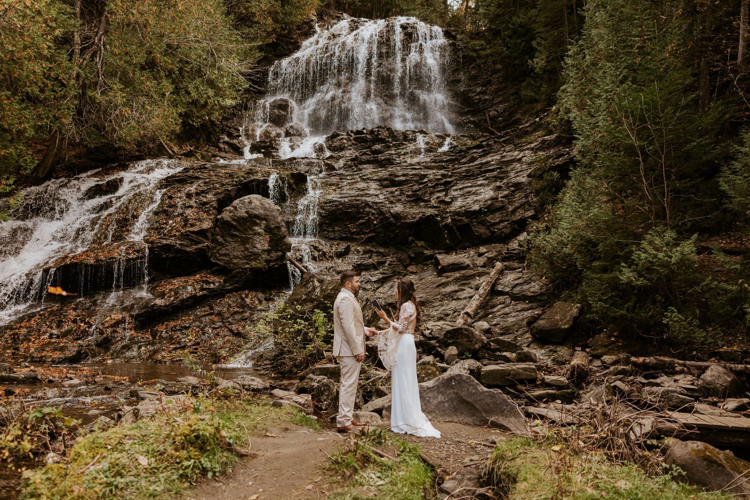 Couple exchanges vows on rocky path in front of a majestic waterfall during an intimate fall elopement.
