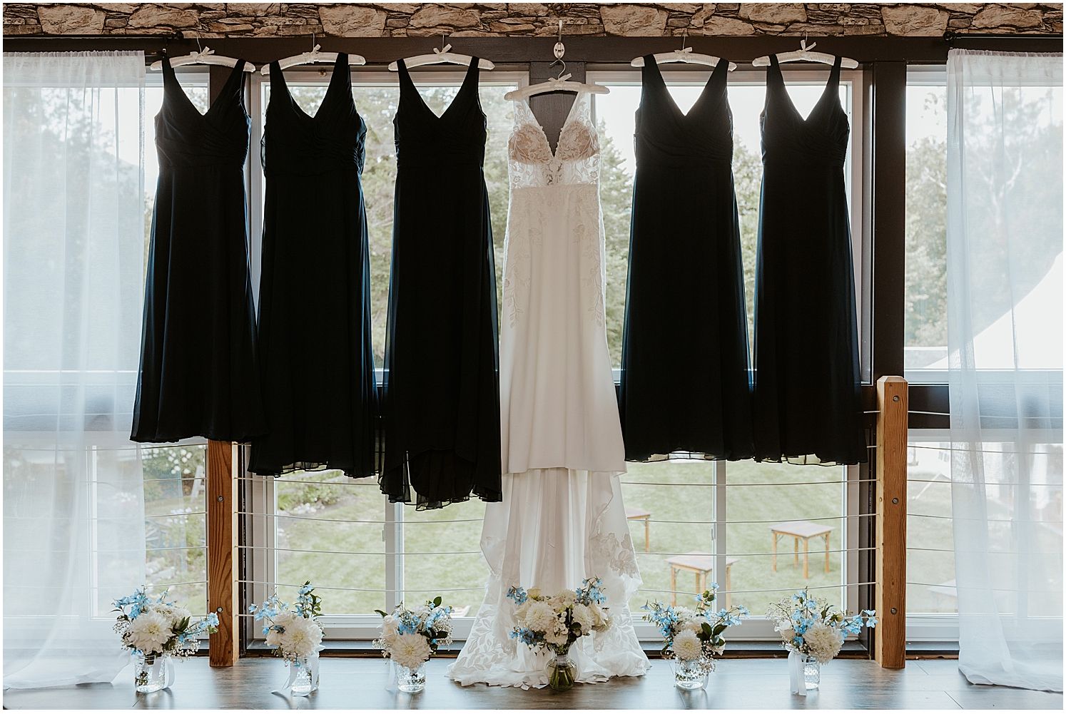 Wedding party attire featuring black bridesmaid dresses and a white wedding gown hanging on a rustic wooden beam.