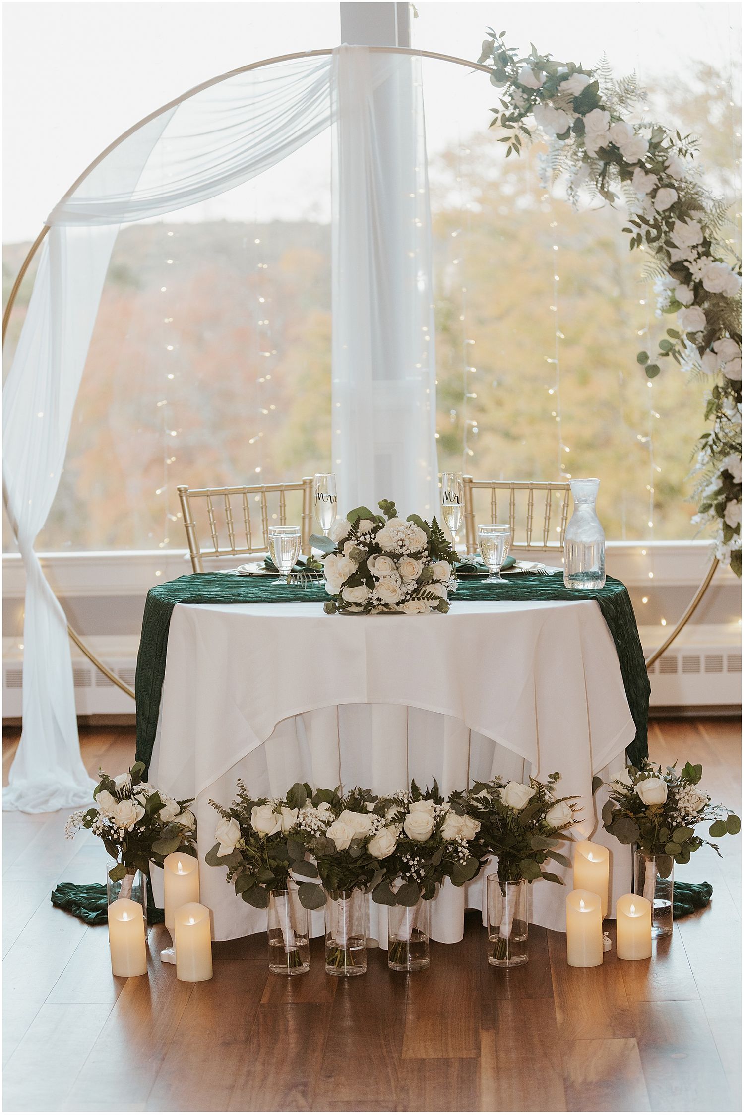 Elegant sweetheart table setup with white linens, green accents, and floral arch decorations.