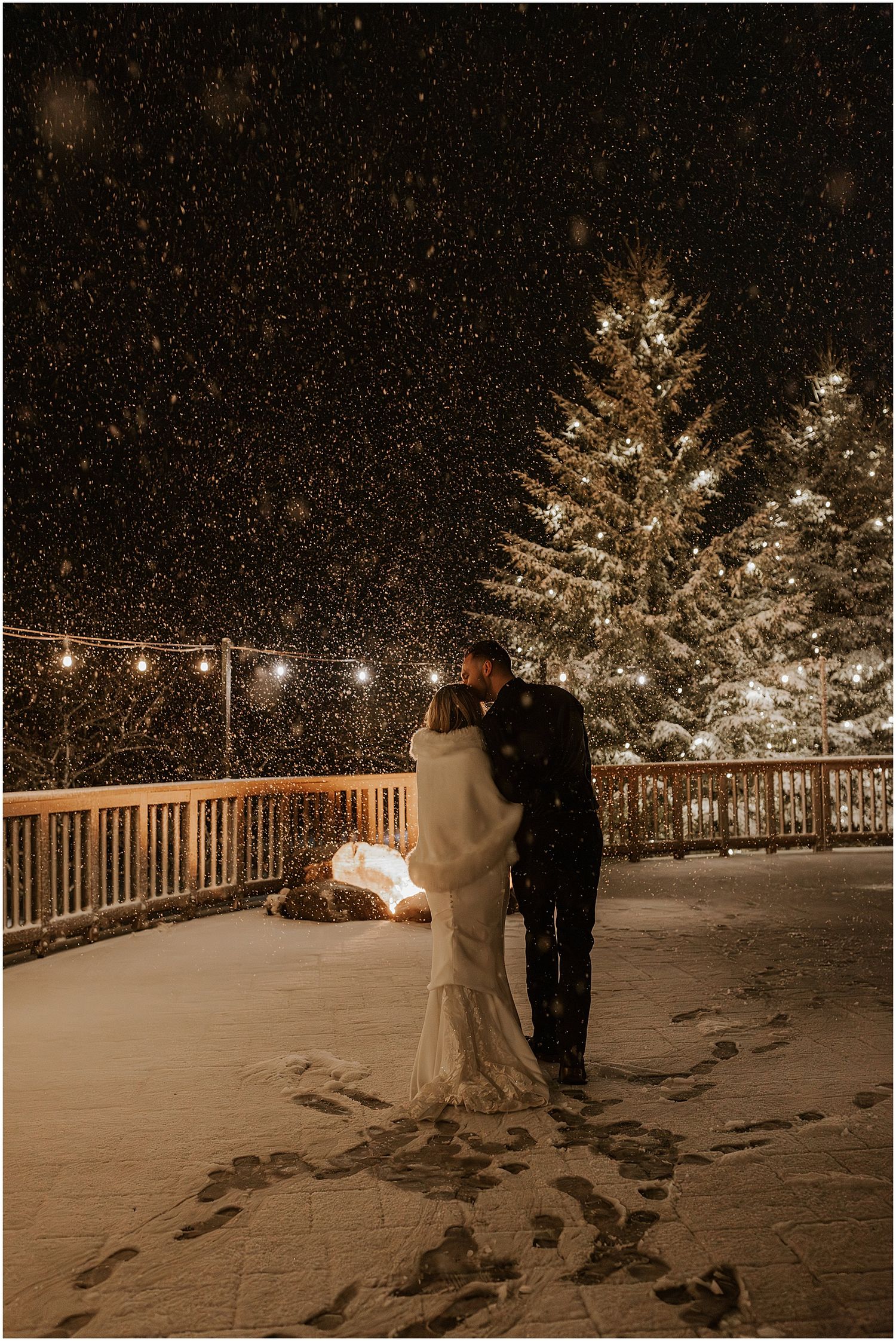 A magical winter wedding moment captured with falling snow and twinkling lights on a snowy deck.