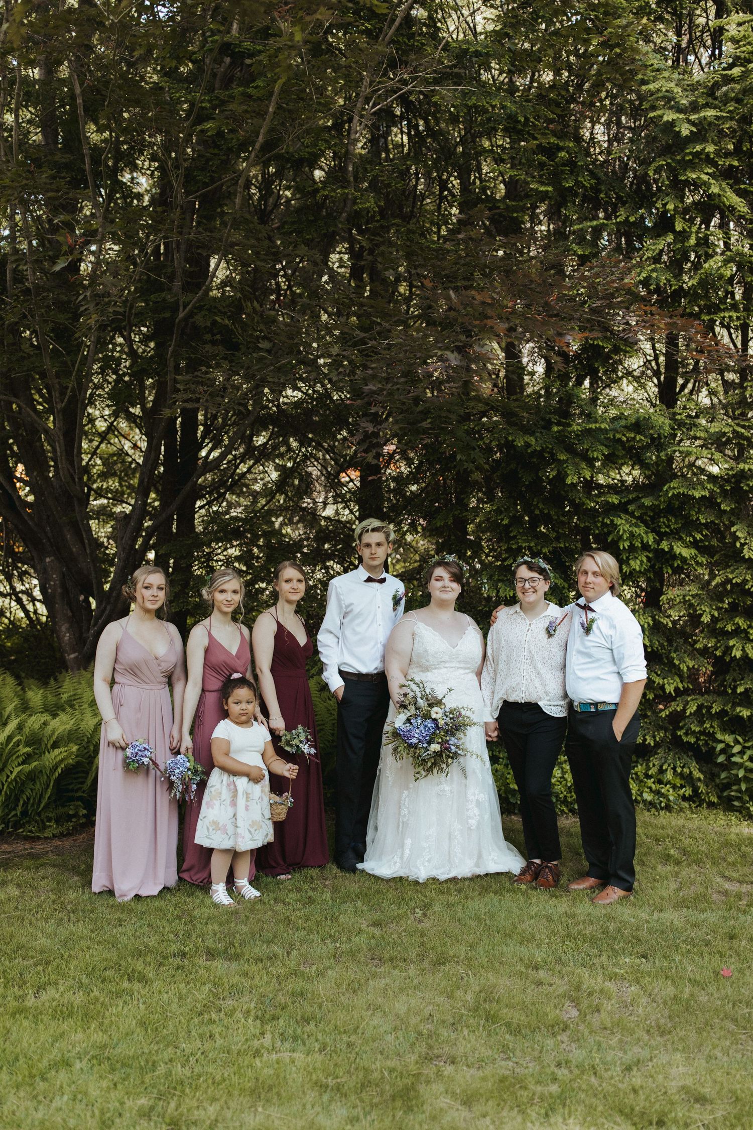 Wedding party poses together for formal group photo in garden setting with trees.