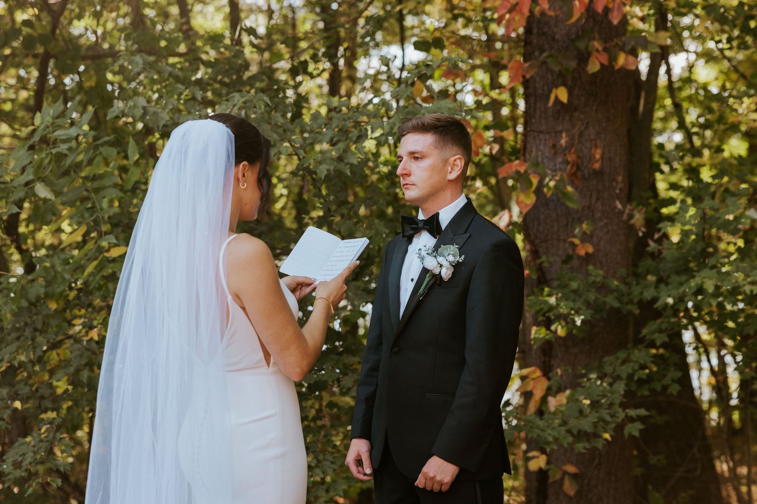 Bride in white gown and veil reads vows from booklet to groom in black tuxedo among autumn trees.