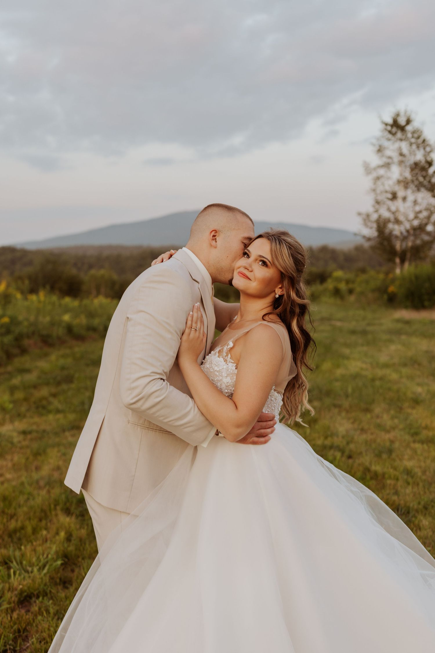 Groom in tan suit kisses bride in white ballgown in green meadow with mountain backdrop at sunset.