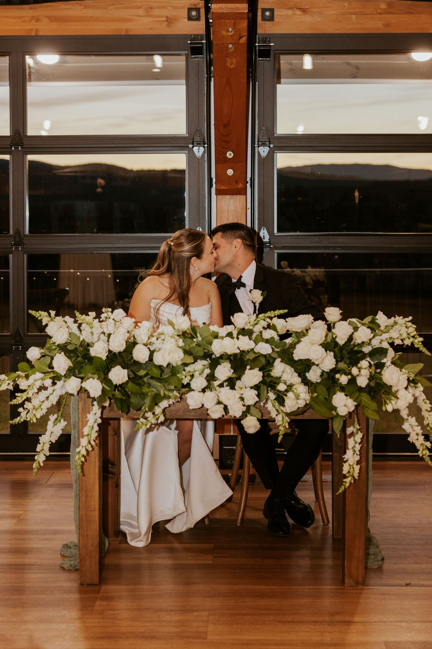 Bride and groom share a kiss at sweetheart table surrounded by white floral decor.