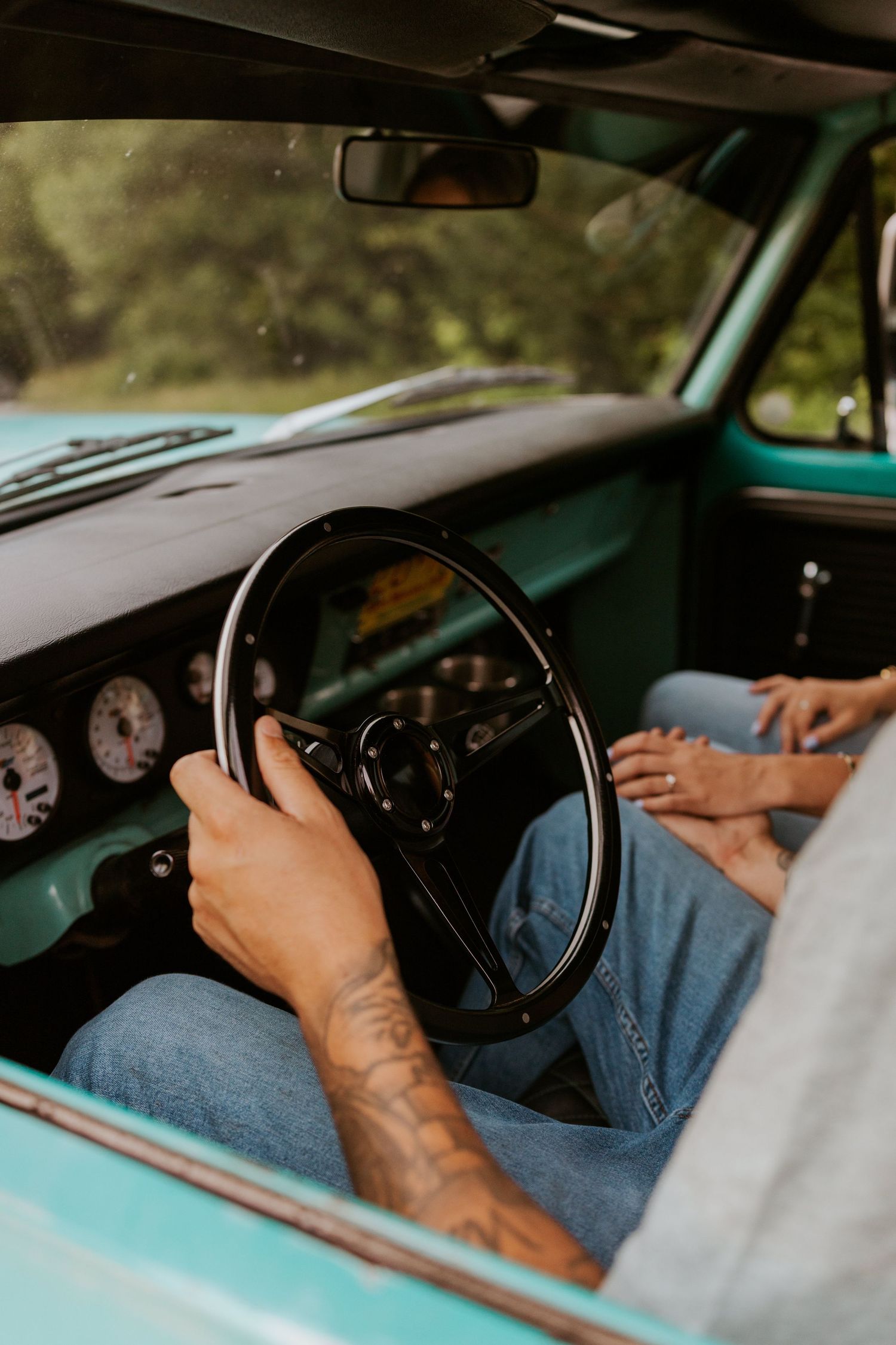 Tattooed man gripping a black steering wheel inside a classic teal pickup truck interior.