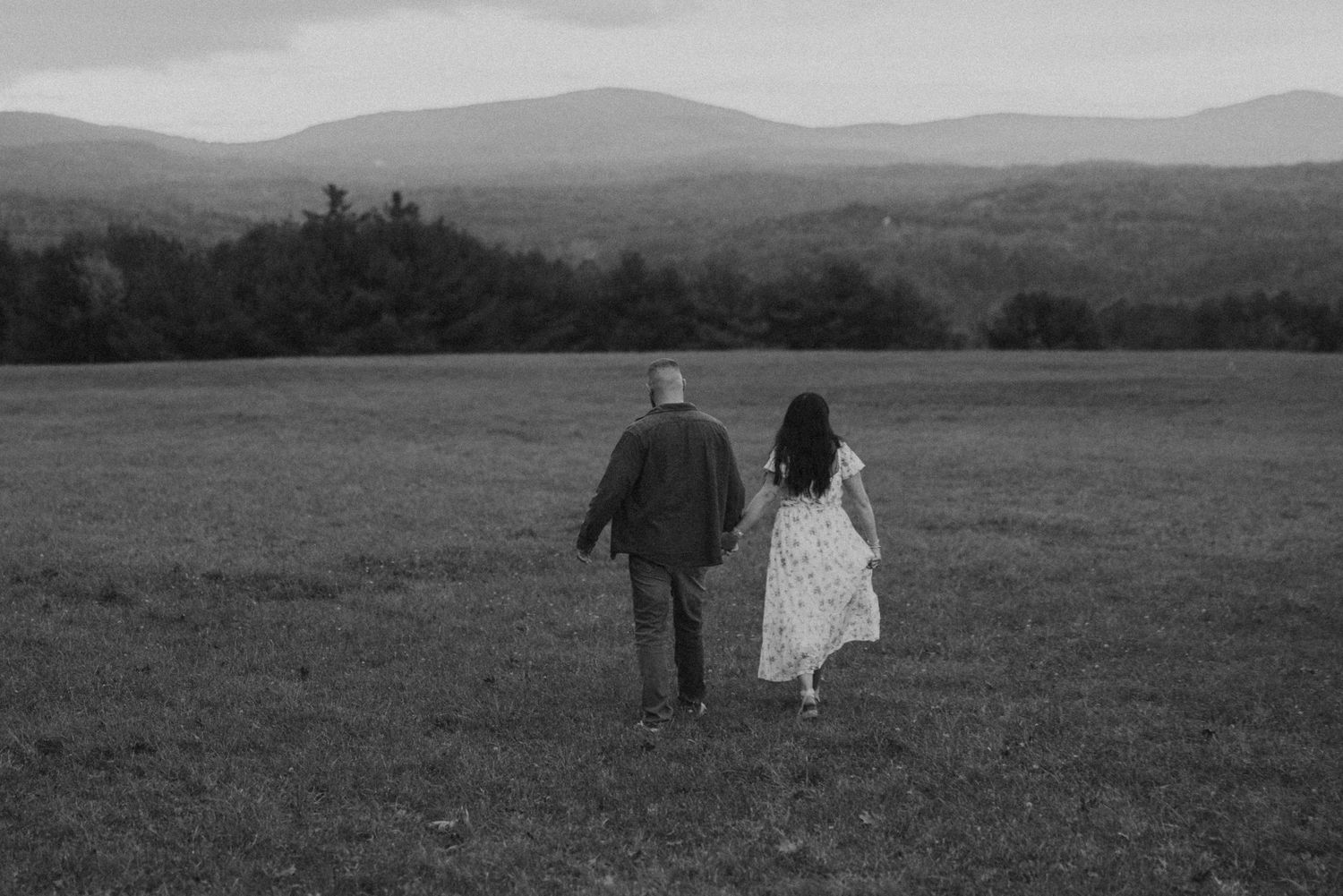 Black and white photo of couple walking hand in hand across open mountain field.