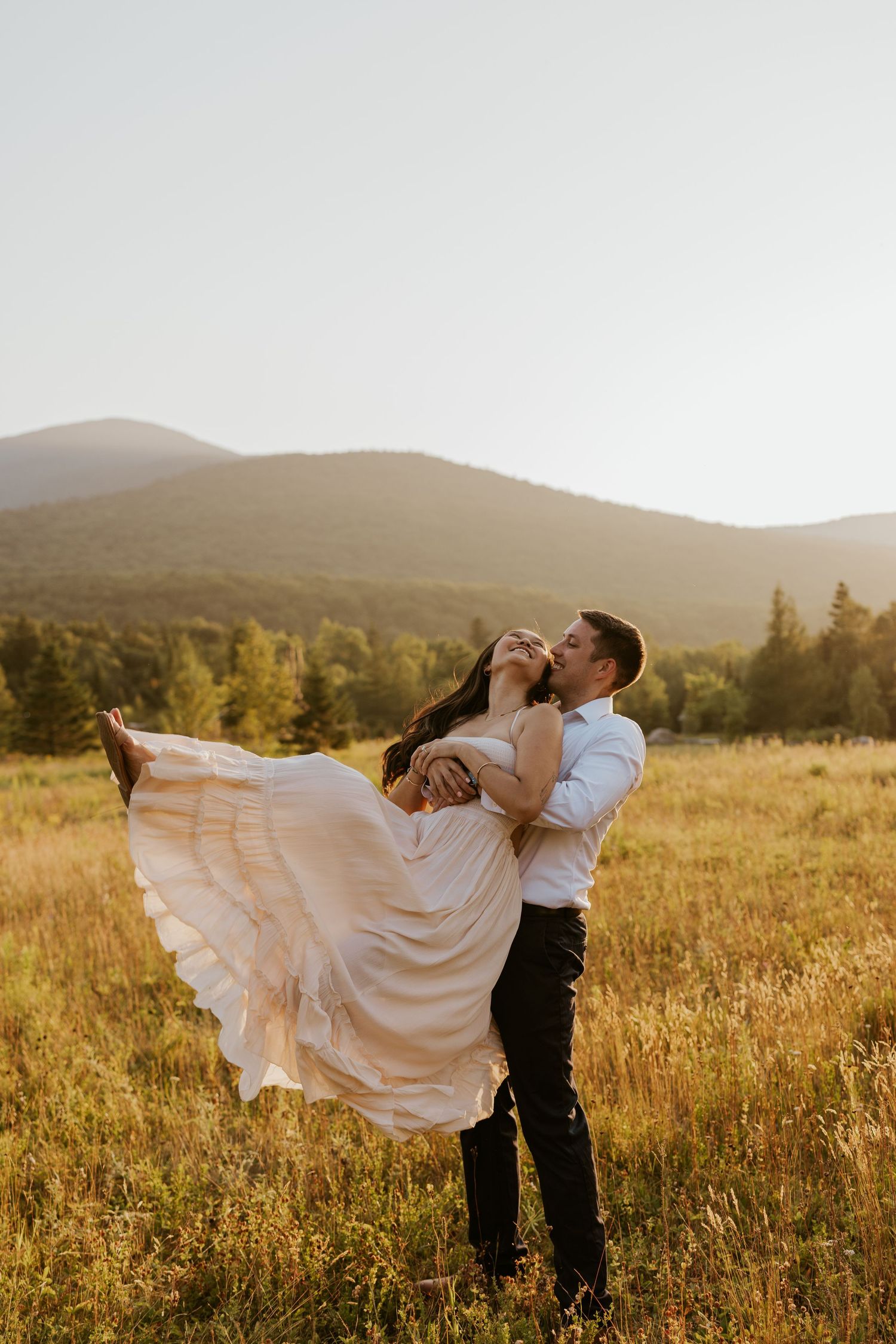 Groom lifts bride in blush dress joyfully in sun-drenched mountain meadow.