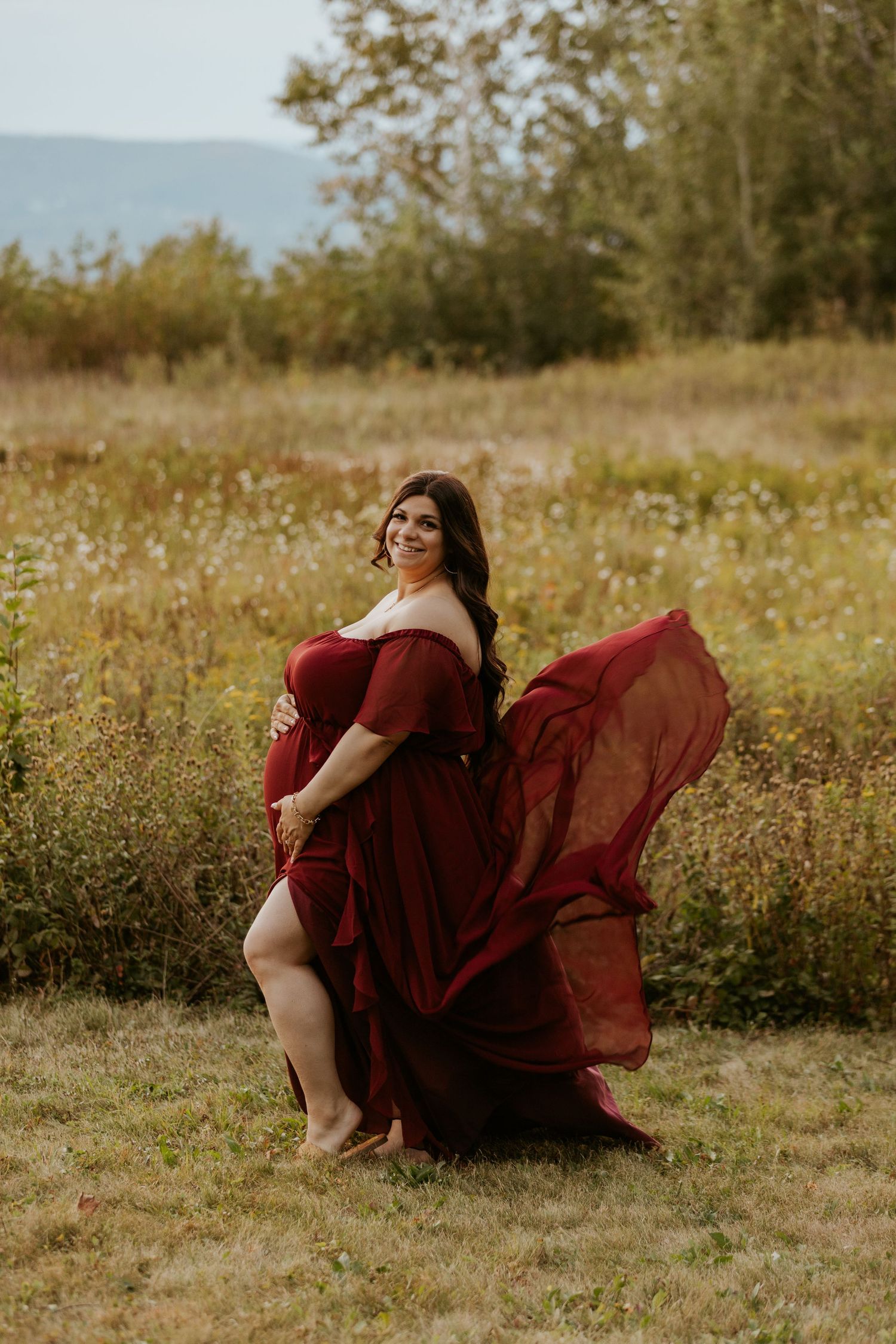 Pregnant woman in flowing red maternity gown posing in wildflower meadow with mountains behind her.