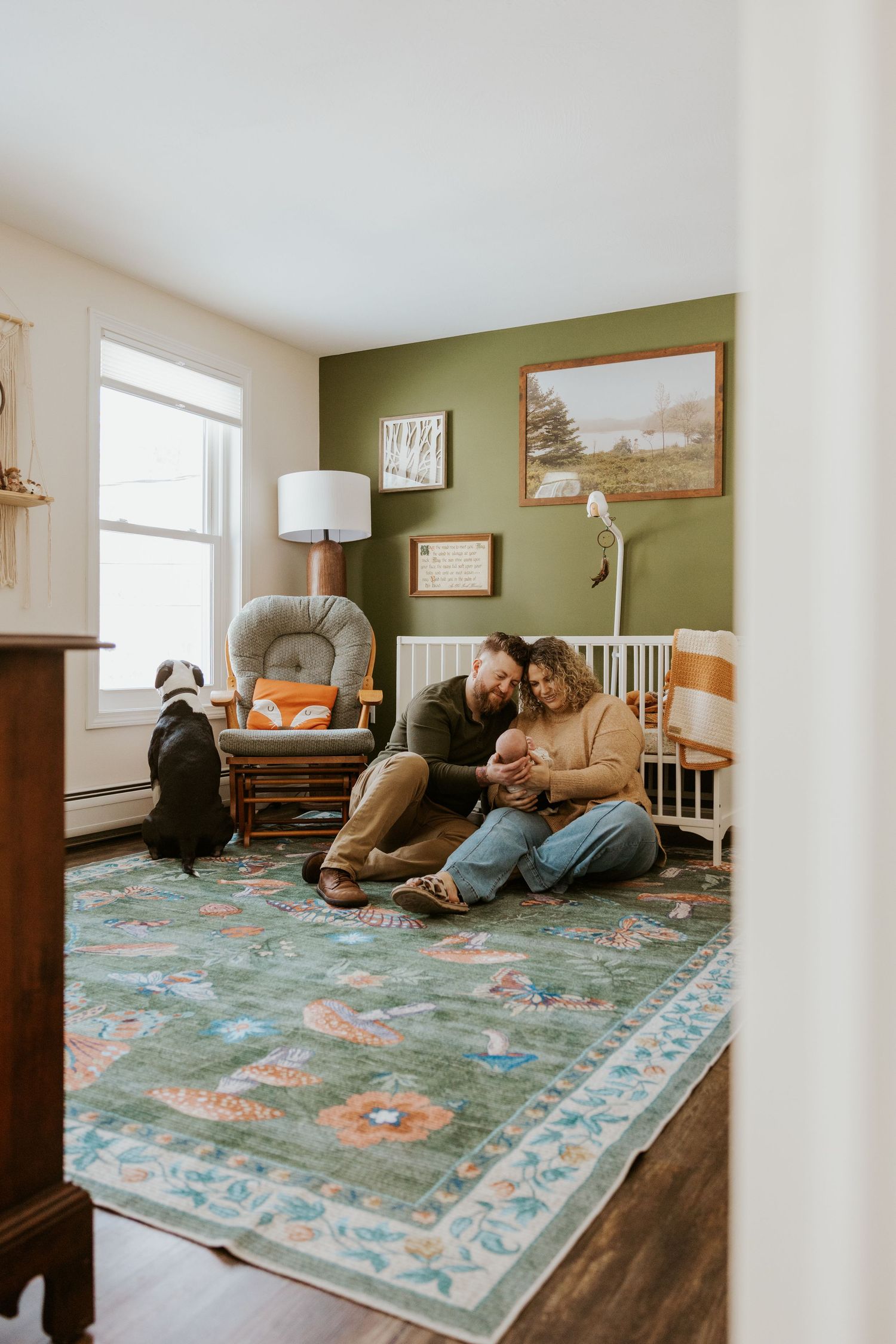 New parents sit on nursery floor with newborn baby beside white crib and green accent wall.