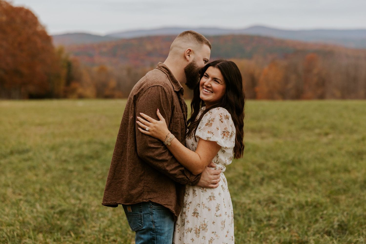 Couple embracing and laughing together in a green field surrounded by vibrant autumn mountain scenery.