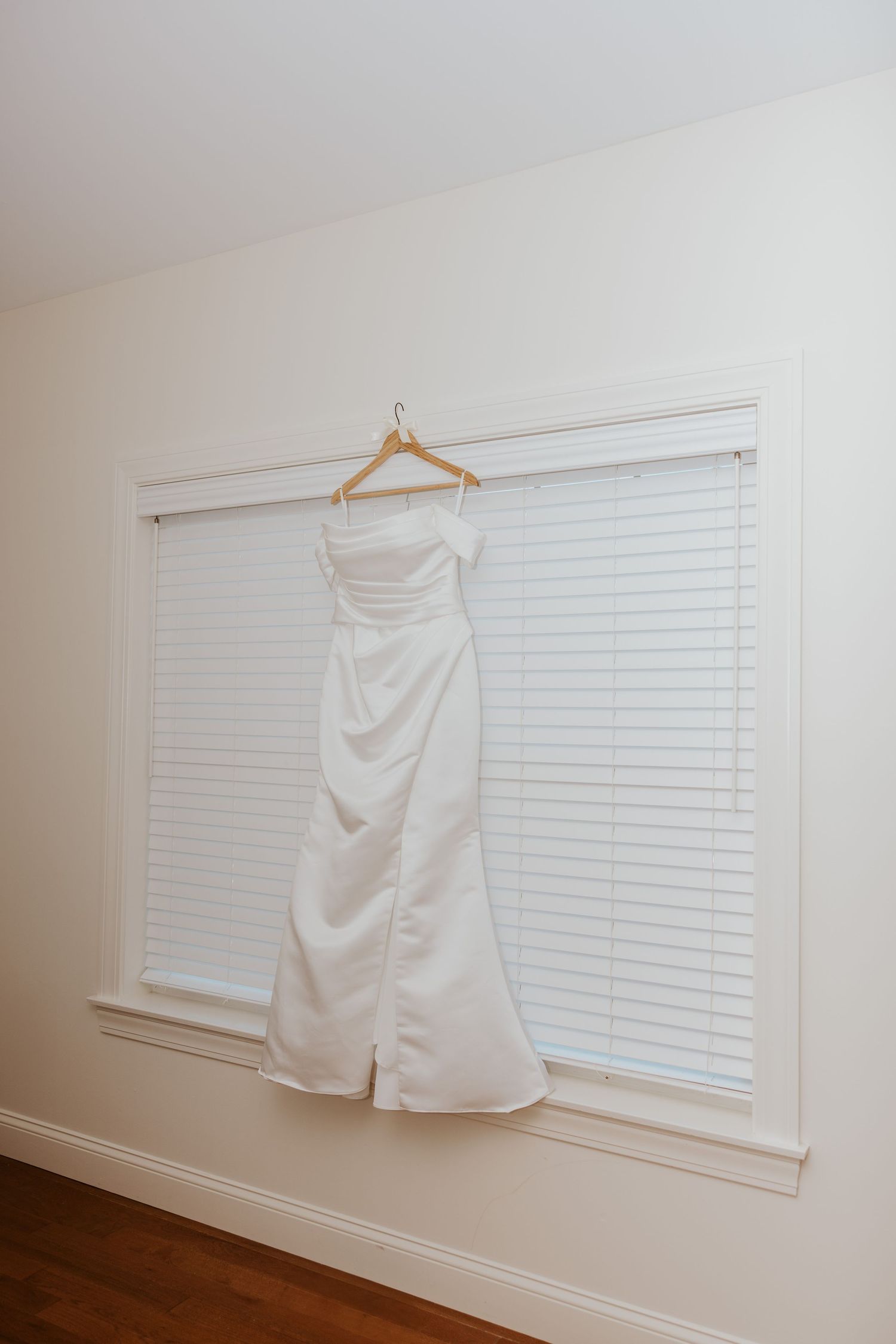 Elegant white off-shoulder satin wedding gown hanging on a wooden hanger in front of a bright window.