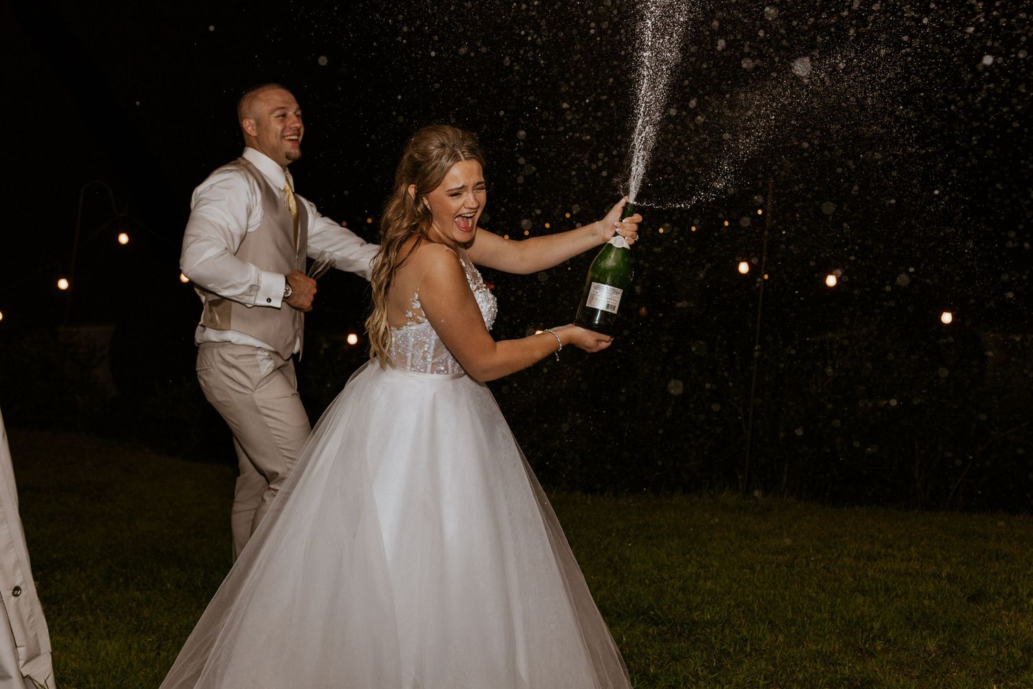 Bride pops champagne bottle outdoors at night as groom watches during wedding celebration.