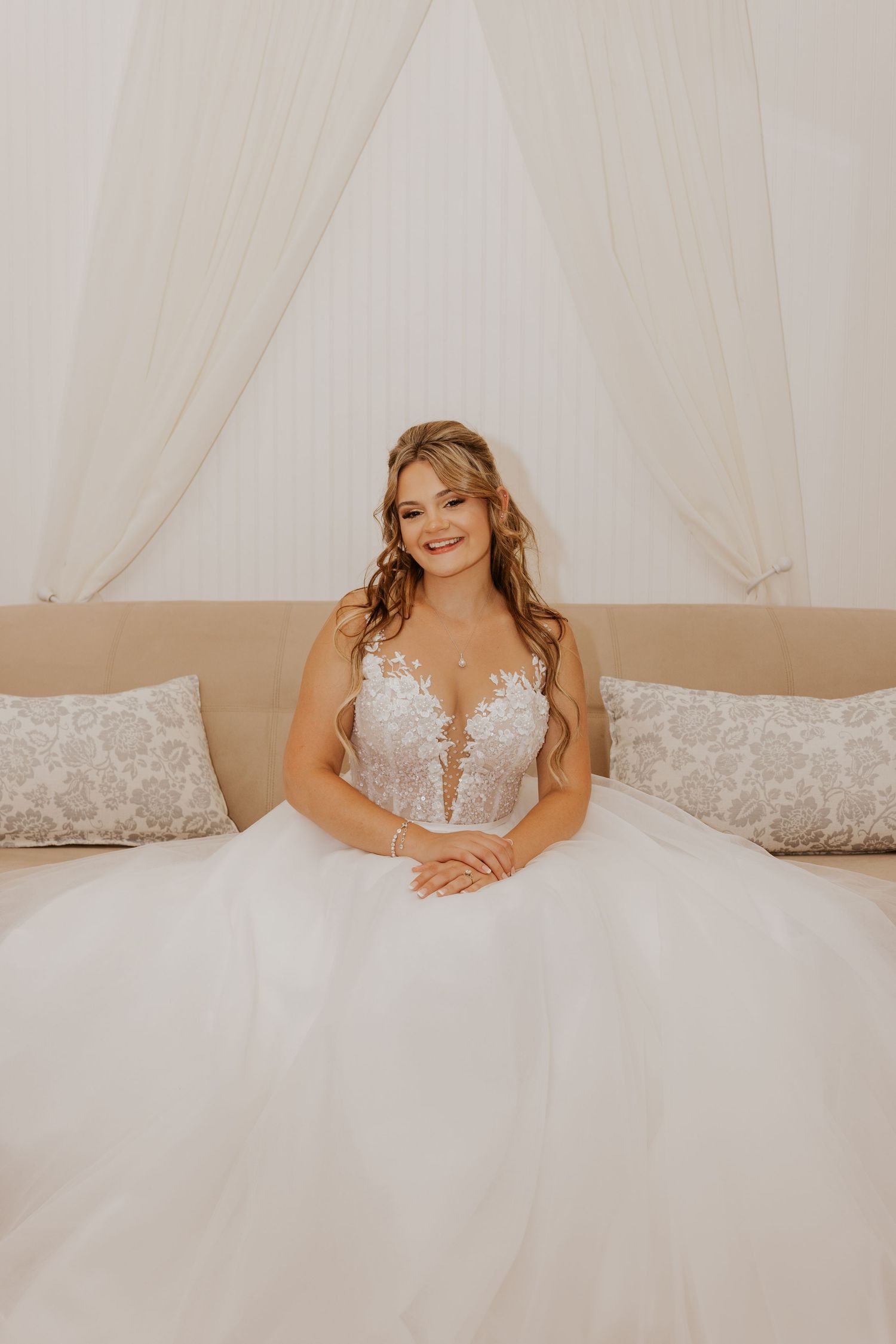 Beautiful bride in a floral lace ballgown sitting on a cream sofa surrounded by white draping.