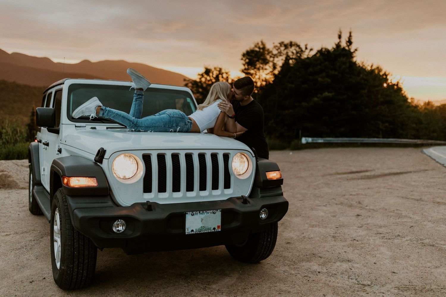 Paige is laying across the hood of her jeep as Justin stands beside her and ksises her at sunset in the White Mountains