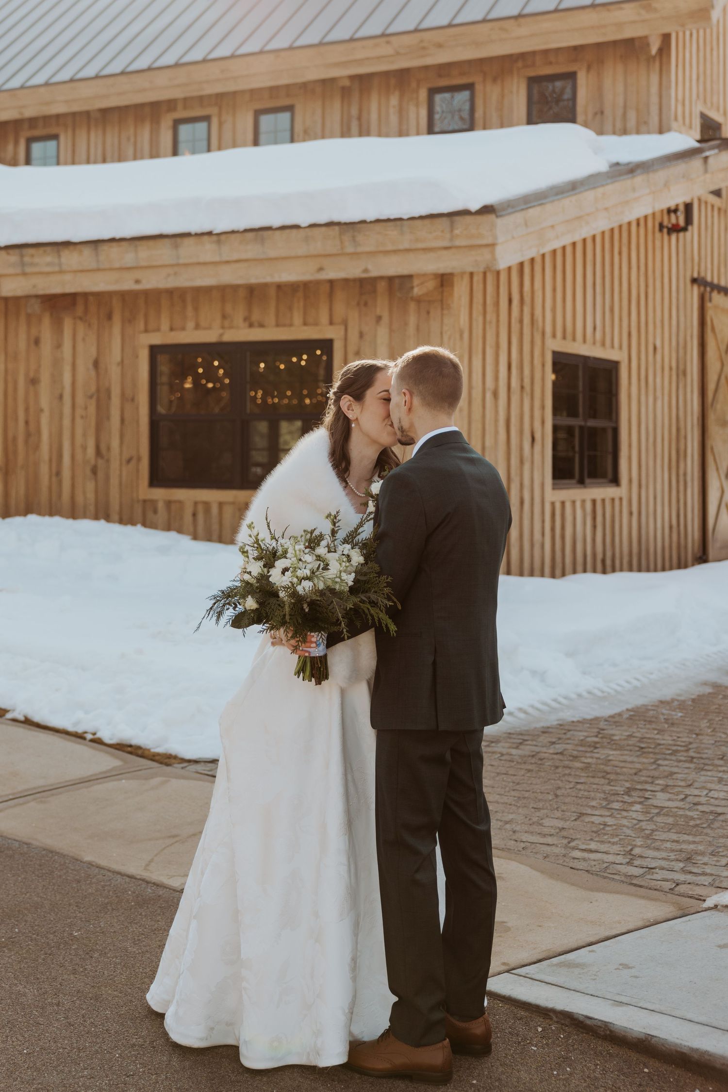A couple in wedding attire share a romantic moment outside a rustic wooden barn in the snow.