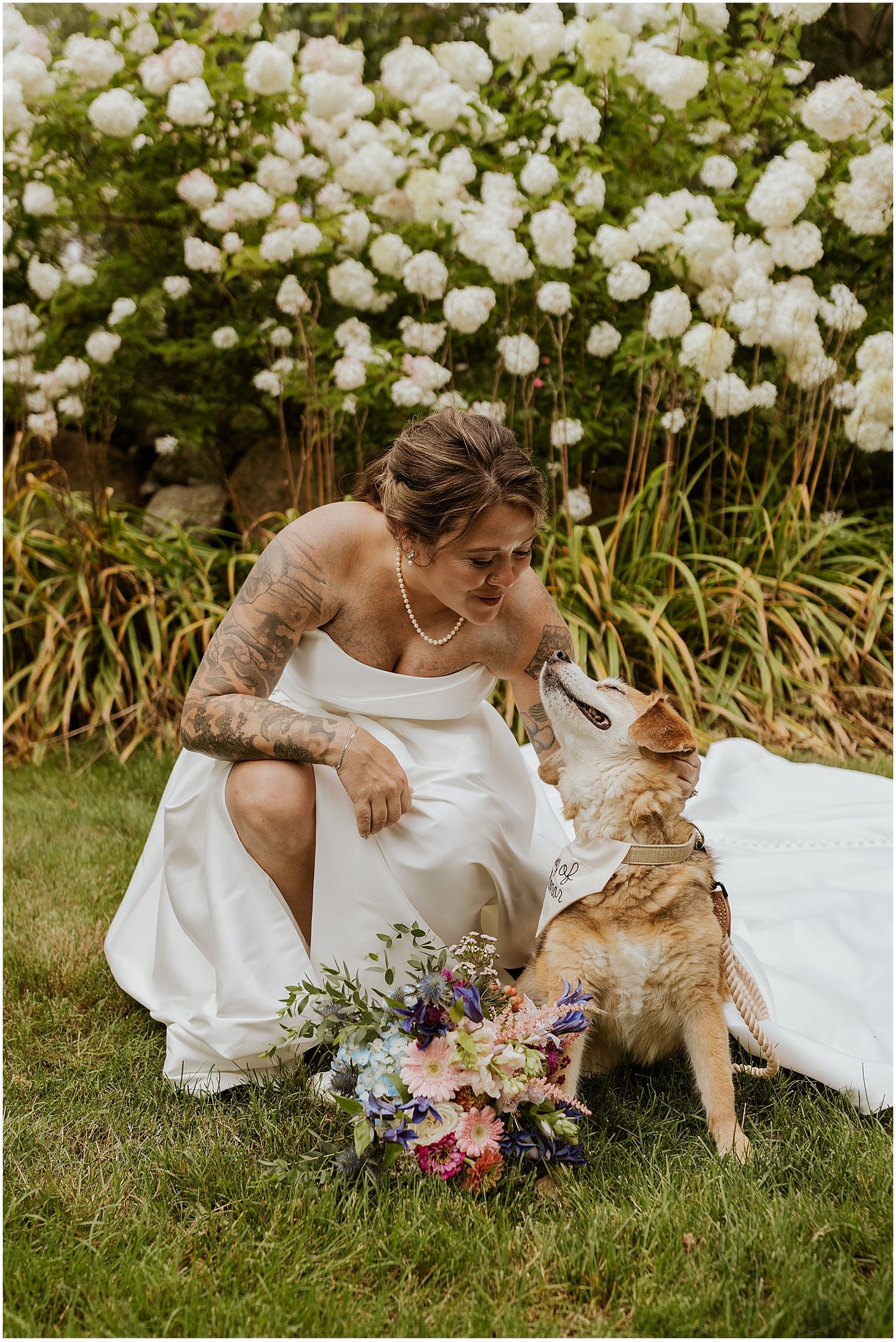 A bride in a white dress sits with a dog next to white flowers and a colorful bouquet in a garden setting.