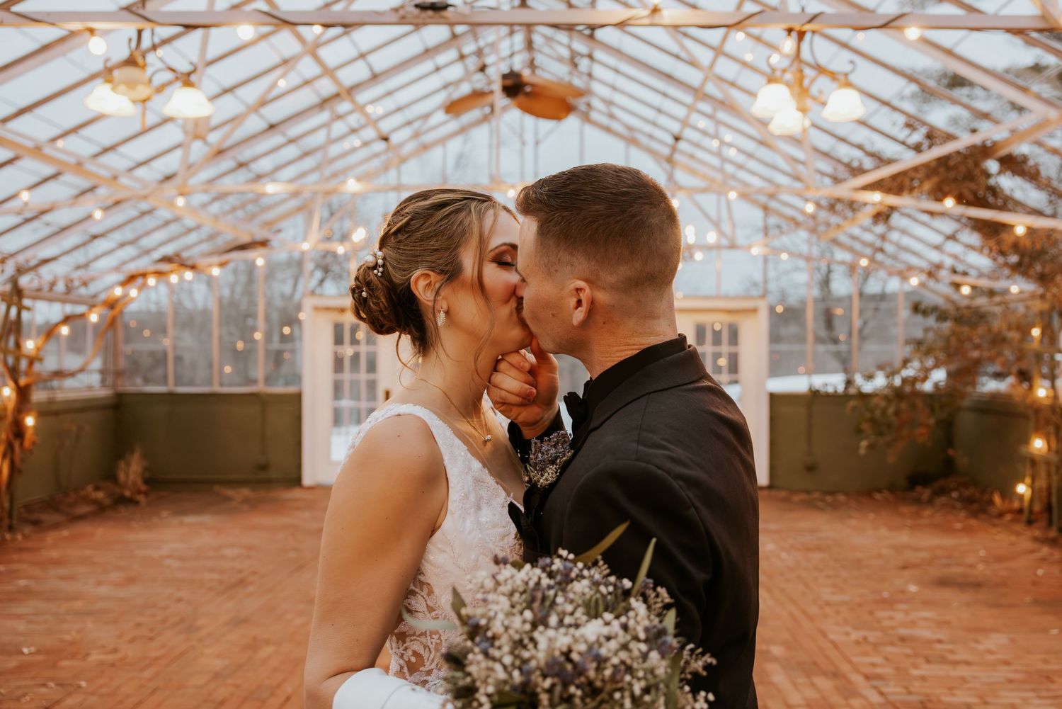 Bride and groom sharing first kiss inside romantic greenhouse venue adorned with string lights.