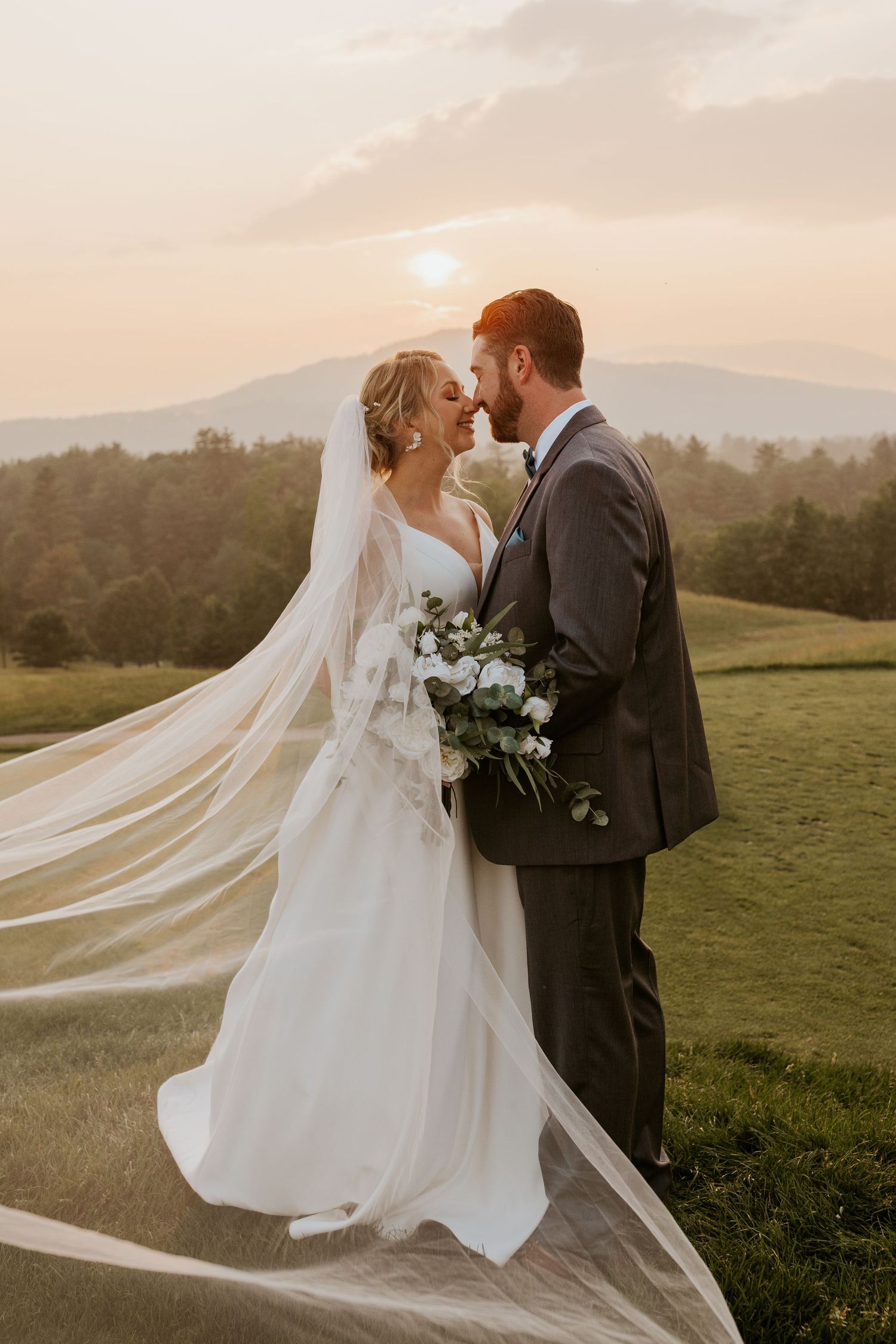 Bride and groom share romantic sunset portrait with flowing veil and mountain landscape backdrop.