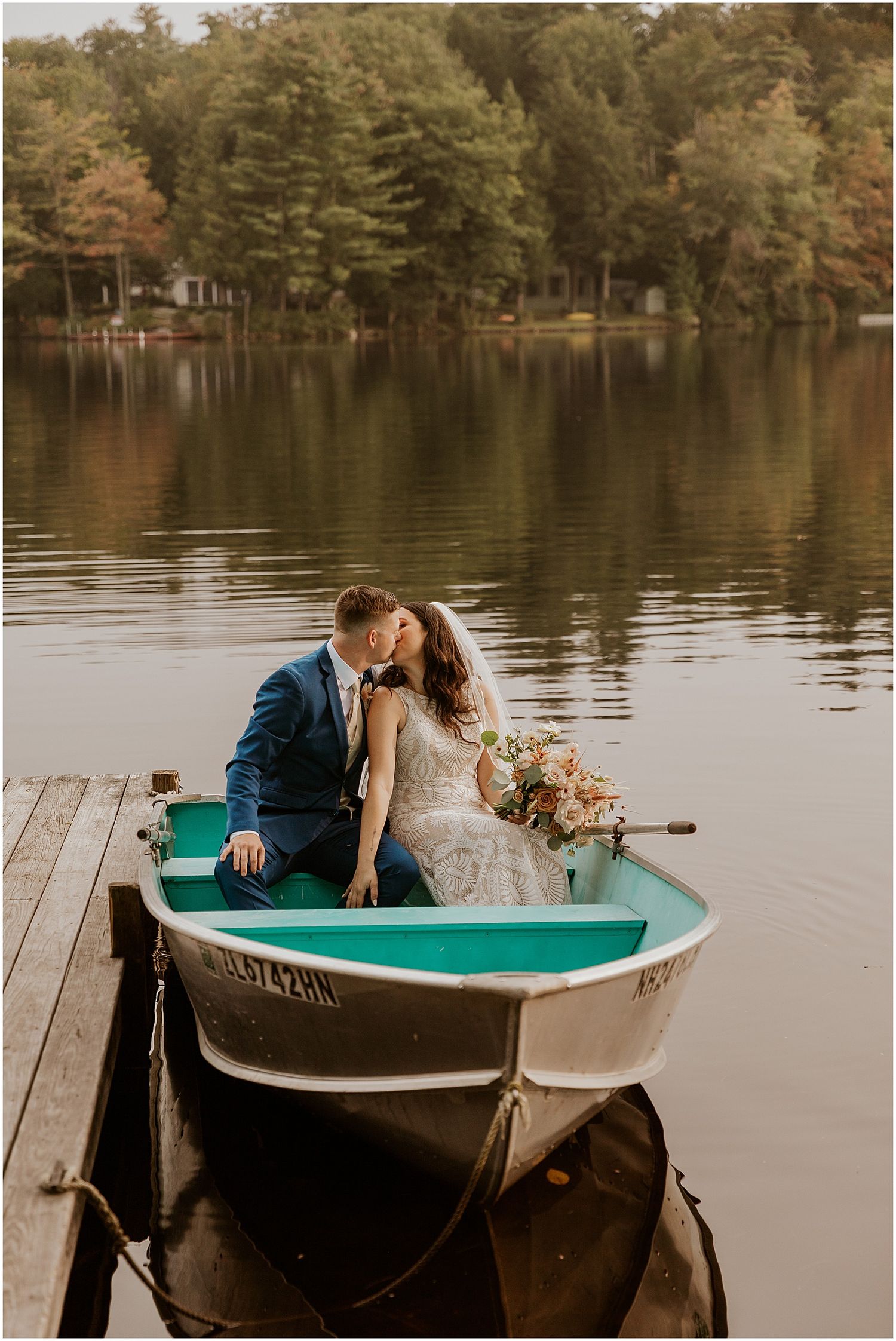 A romantic couple sits in a turquoise rowboat on a calm lake surrounded by autumn trees.