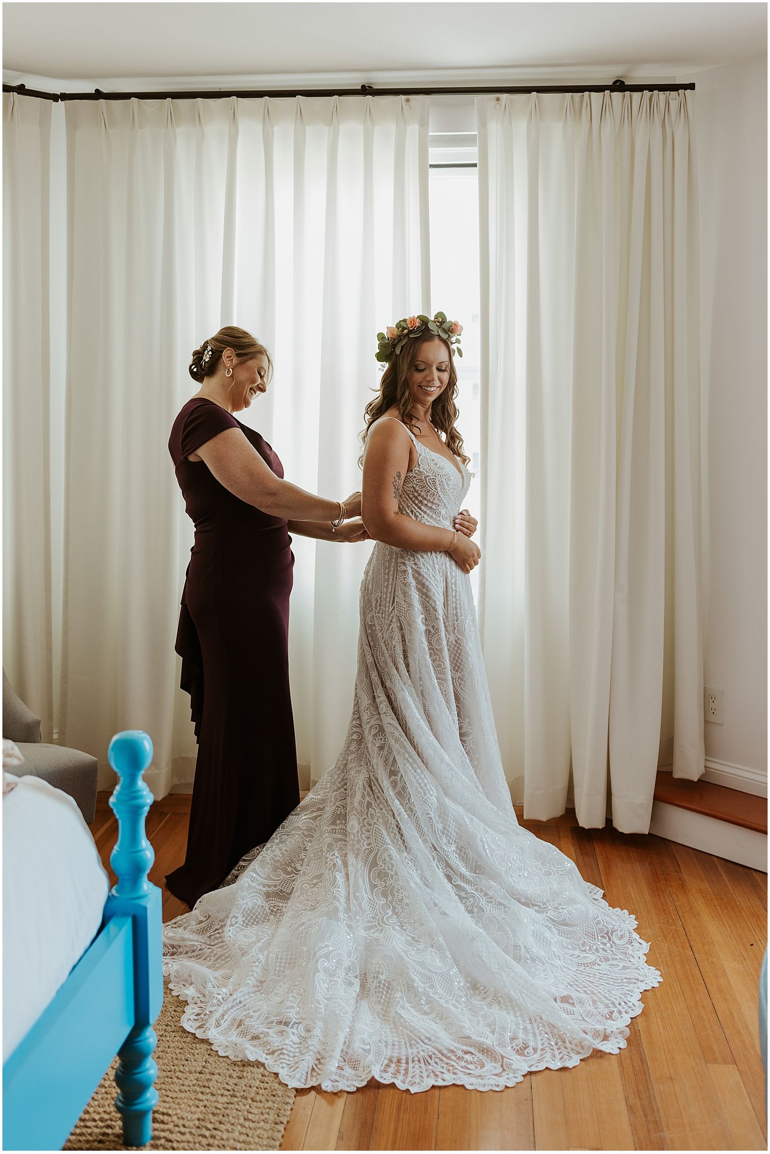 A lace wedding dress with long train being adjusted in a sunlit room with white curtains.