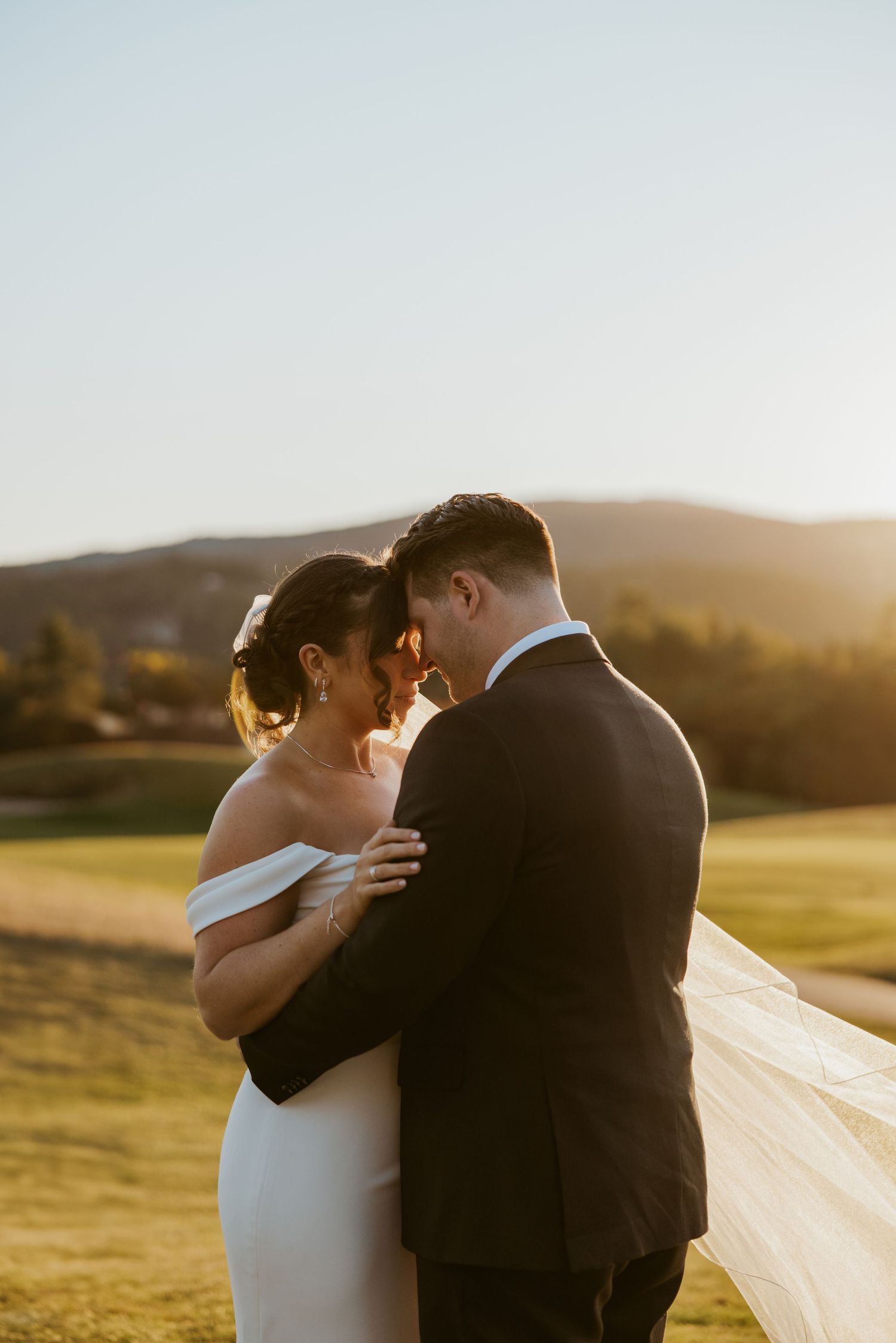 Bride and groom share an intimate moment at golden hour with rolling hills and a flowing veil in the background.