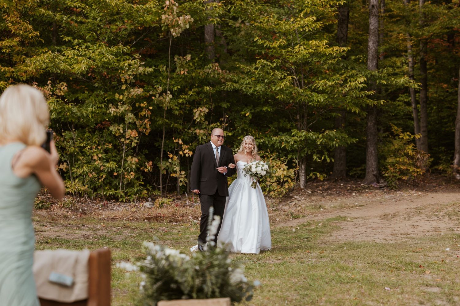 Bride and groom walking hand in hand through a wooded outdoor wedding ceremony with autumn foliage backdrop.