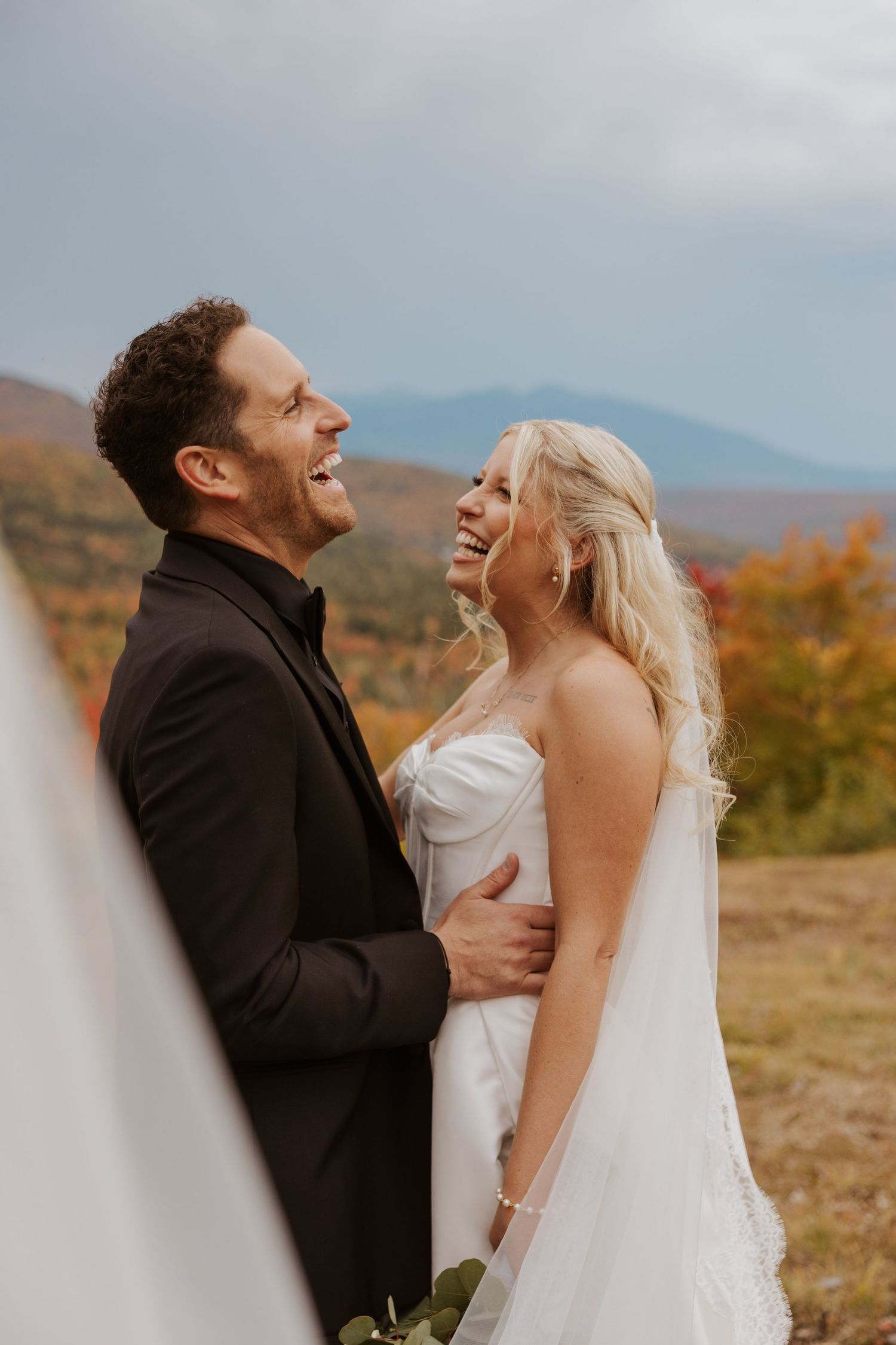 A laughing bride and groom share a joyful moment outdoors with colorful fall foliage behind them.
