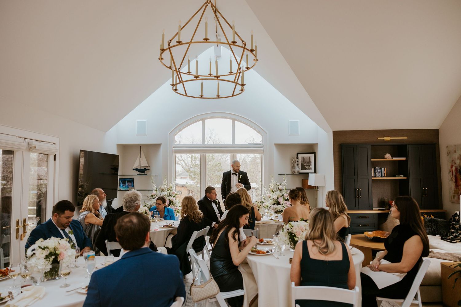 Wedding guests raise their glasses in a toast during an elegant indoor reception with rustic chandeliers.