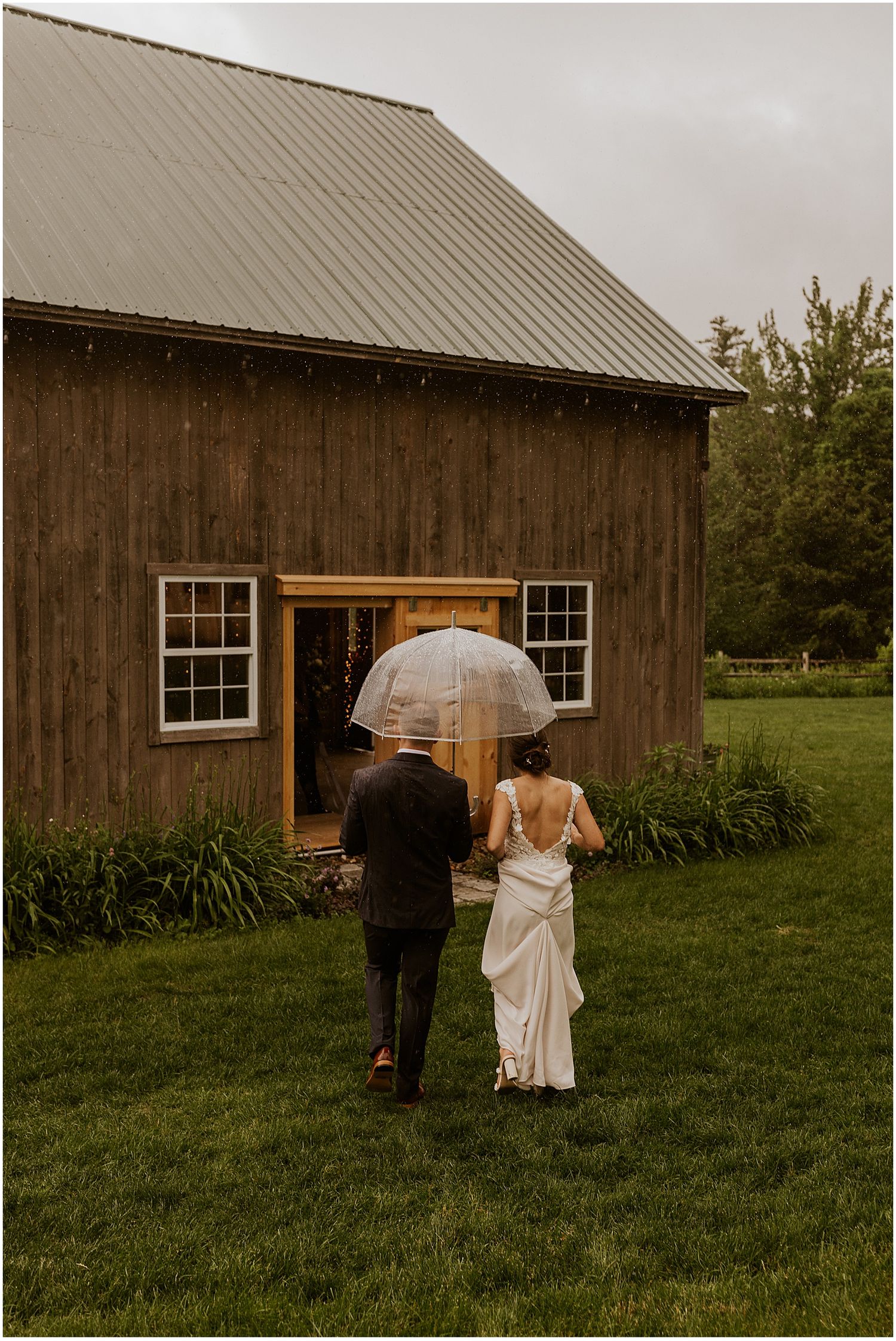 A couple walks along a rustic barn with umbrellas on an overcast day.