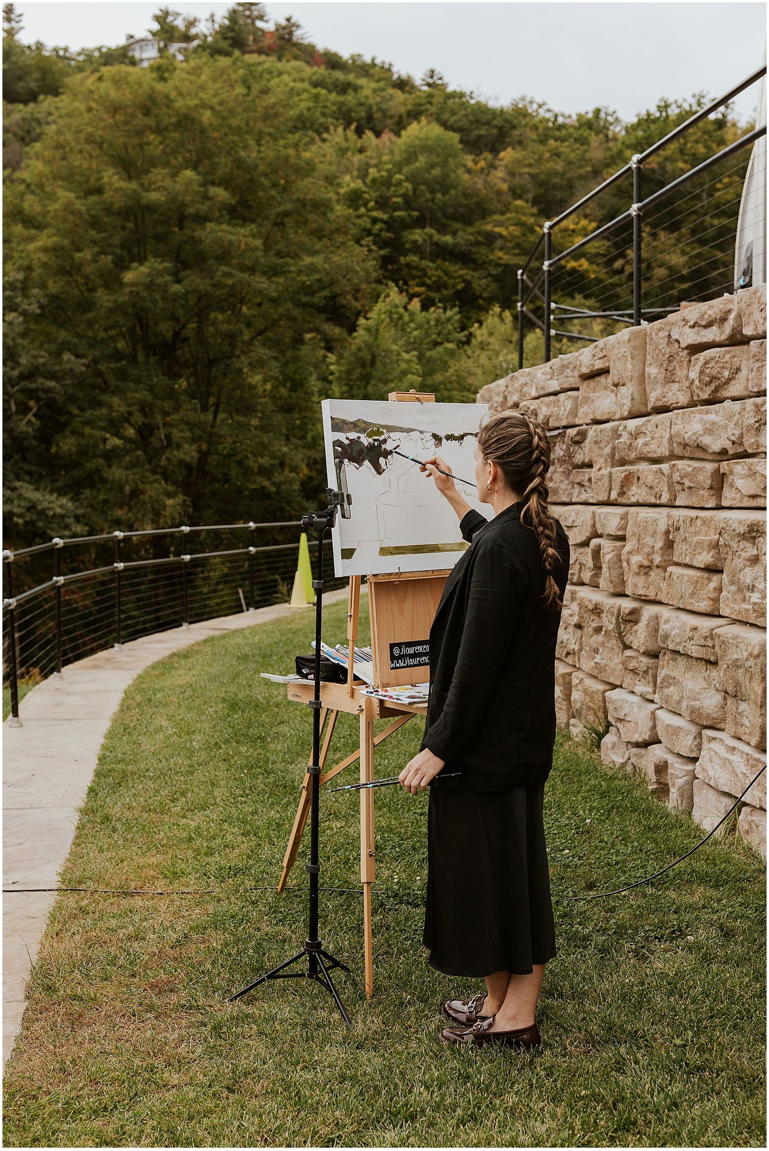Artist painting at an outdoor easel setup along a stone wall path in a natural setting.