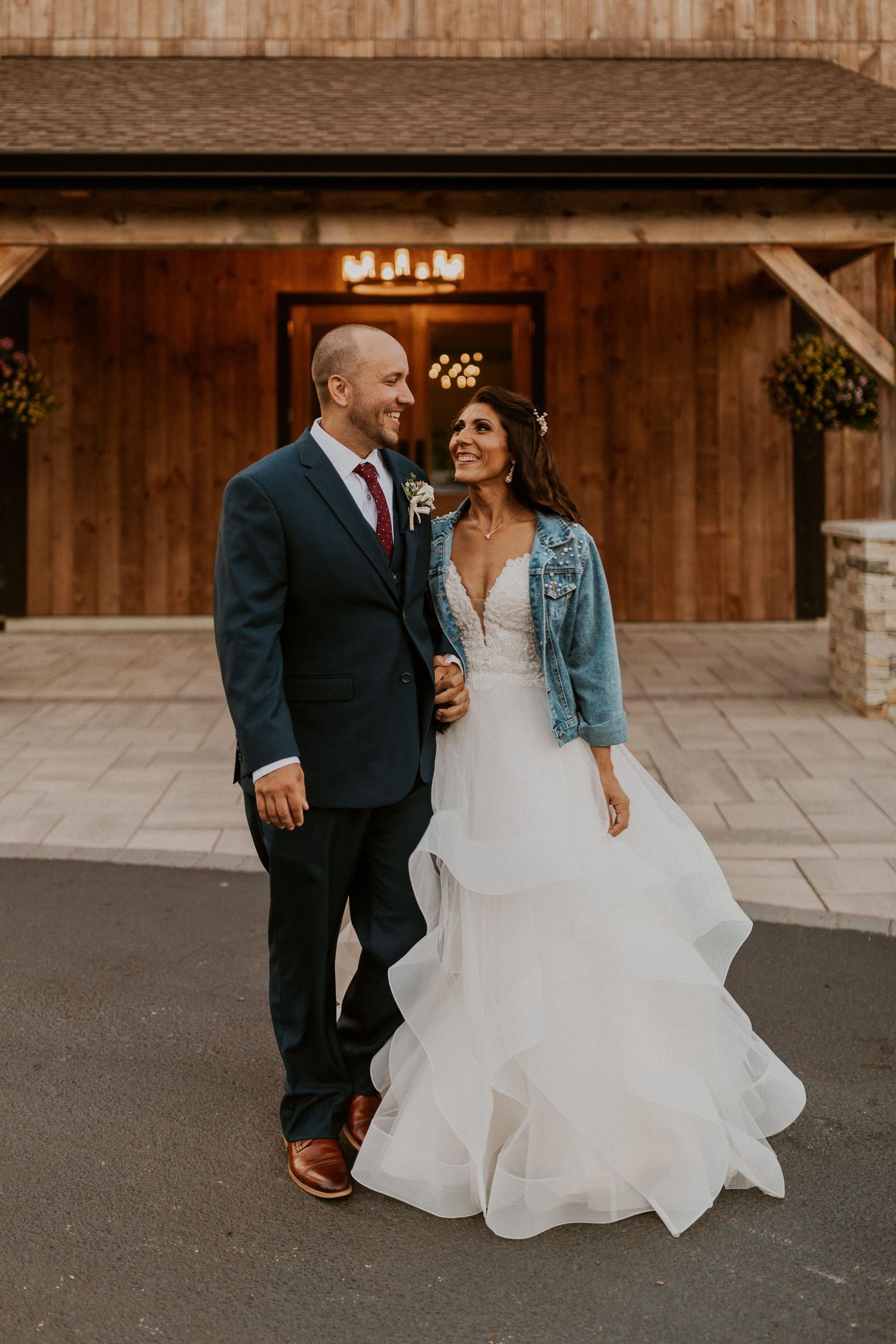 Wedding couple poses playfully outside a rustic barn with the bride wearing a denim jacket.