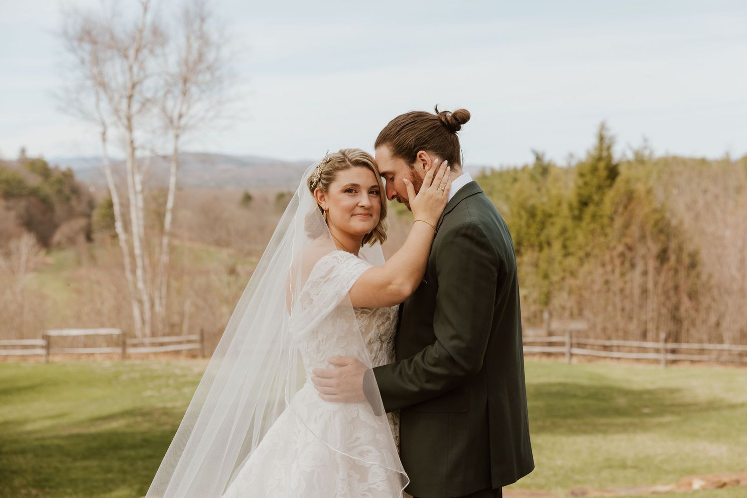 Bride in white gown and veil embraces groom in dark suit outdoors at a wedding with green hills backdrop.