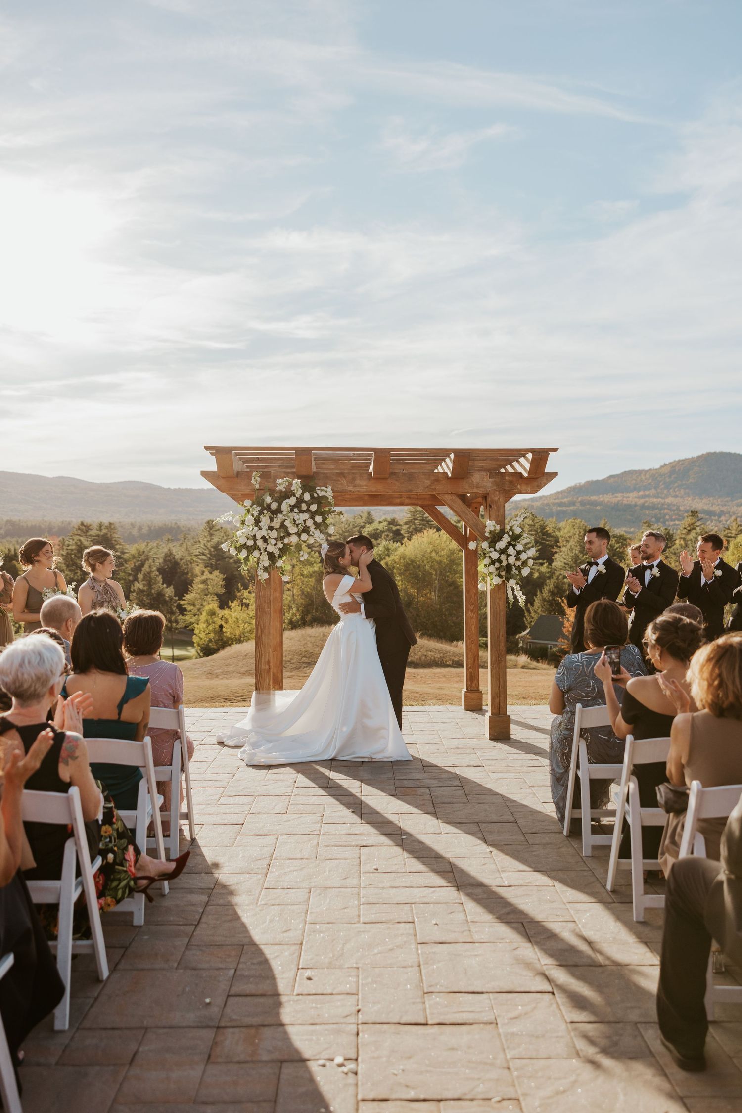 Bride and groom share first kiss at outdoor wedding ceremony under wooden pergola with mountain views.