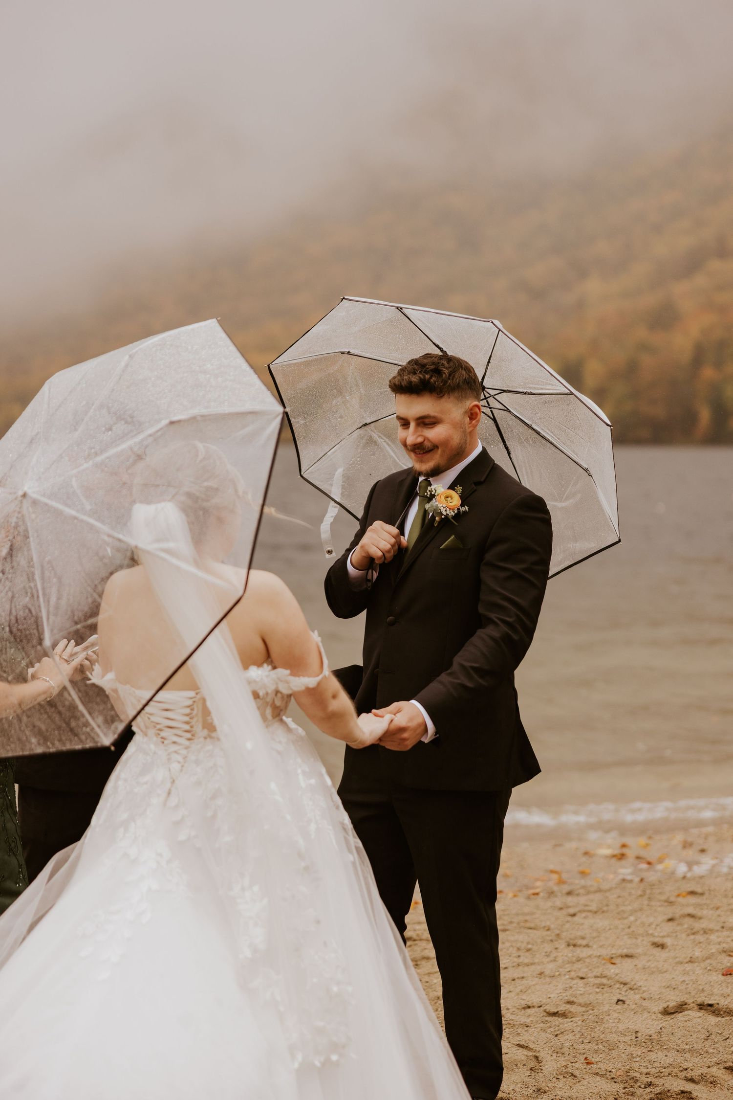 Groom smiles warmly at bride during rainy lakeside wedding ceremony, both holding clear umbrellas.