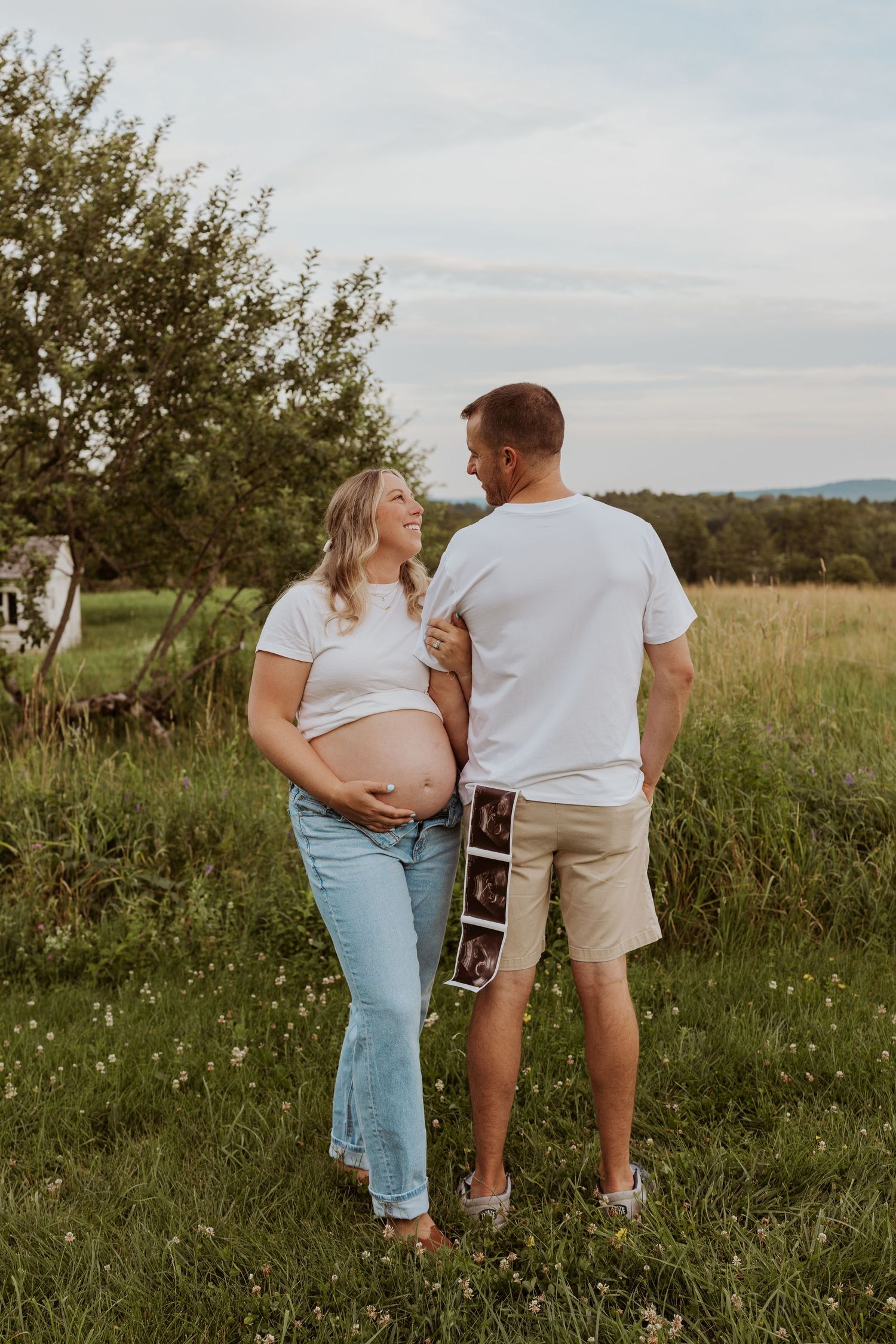 Pregnant woman in white tee holds belly while partner displays ultrasound photos in sunny meadow.
