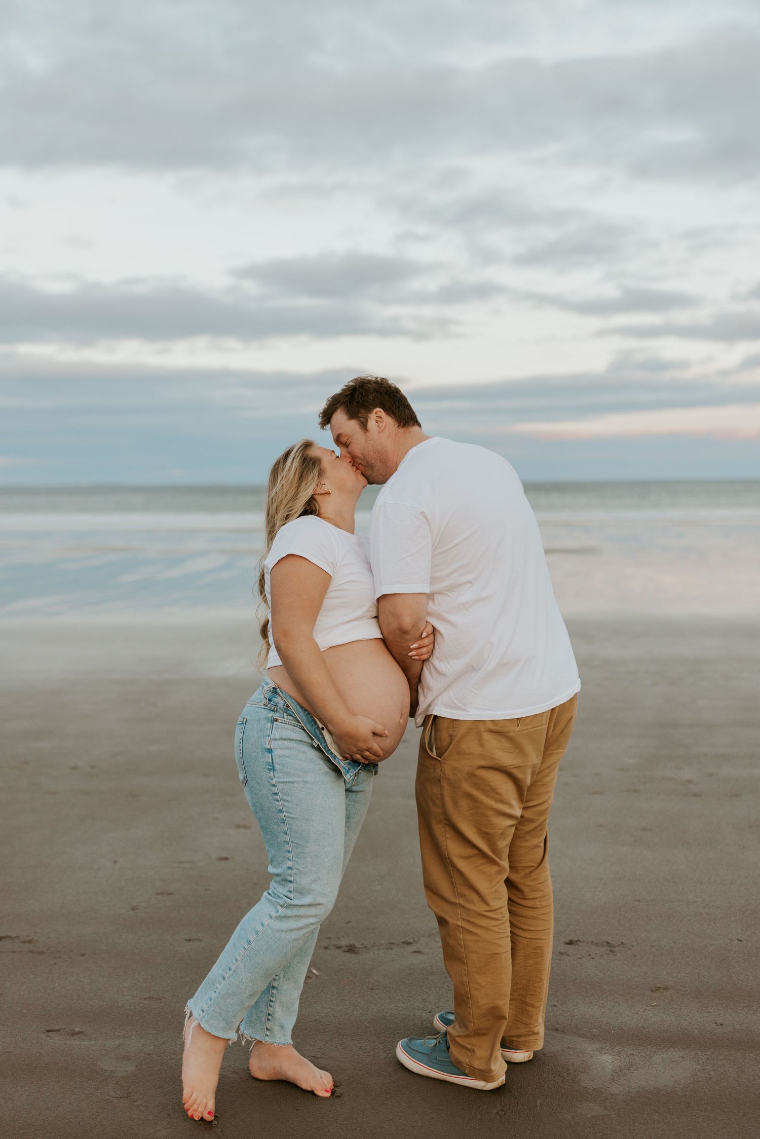 Couple sharing a tender kiss while holding pregnant belly on a cloudy beach.
