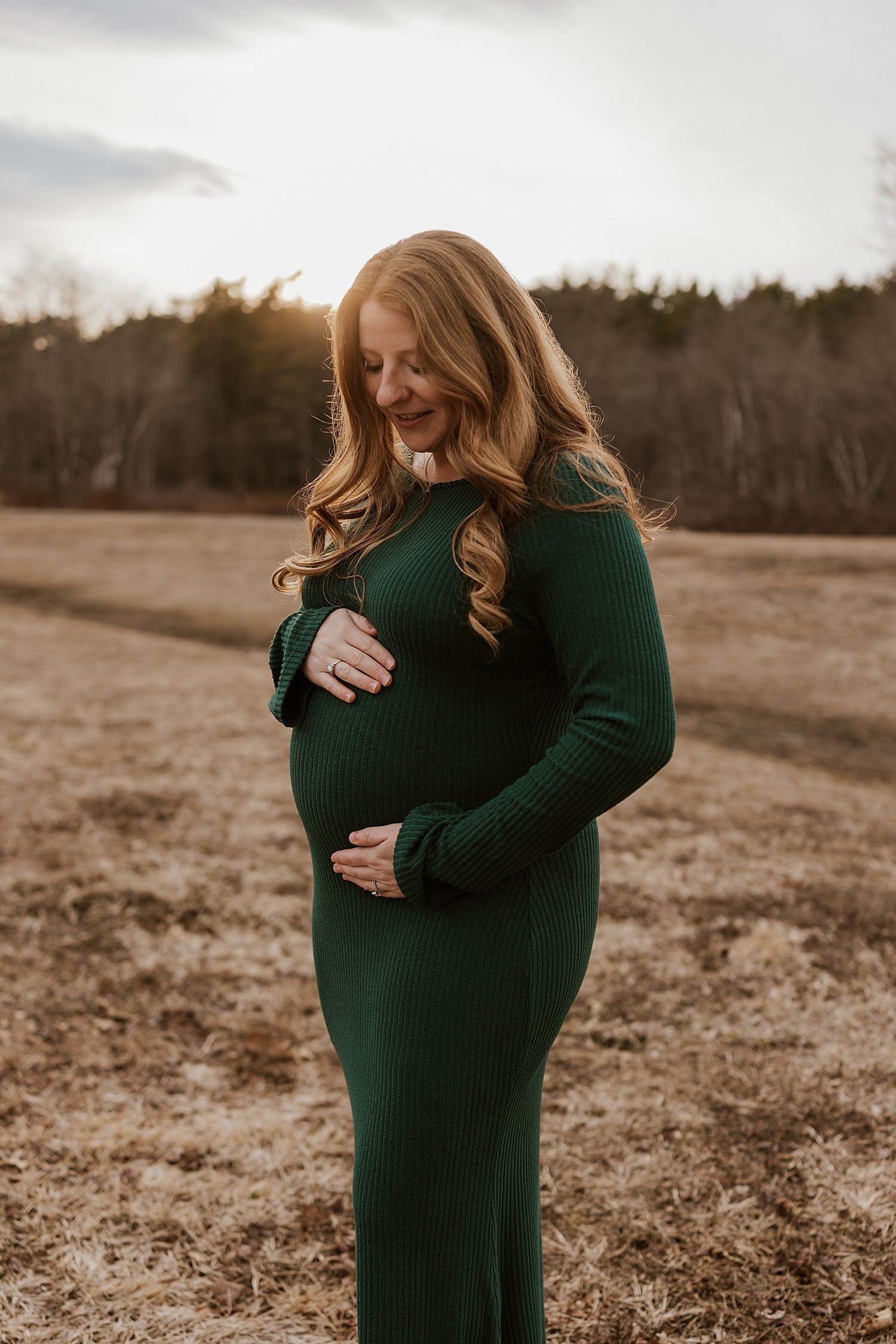 Pregnant woman in green ribbed long-sleeve dress cradling her baby bump in an open field at golden hour.