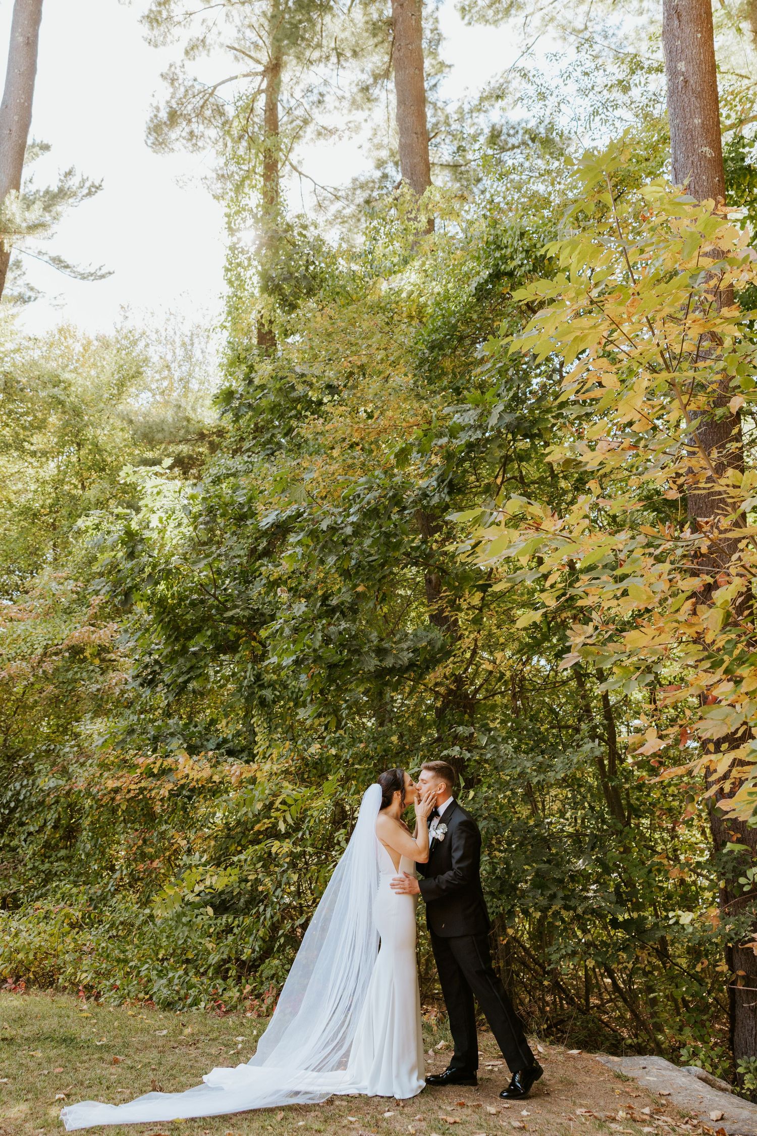 Bride and groom share romantic kiss in wooded outdoor setting with fall foliage backdrop.