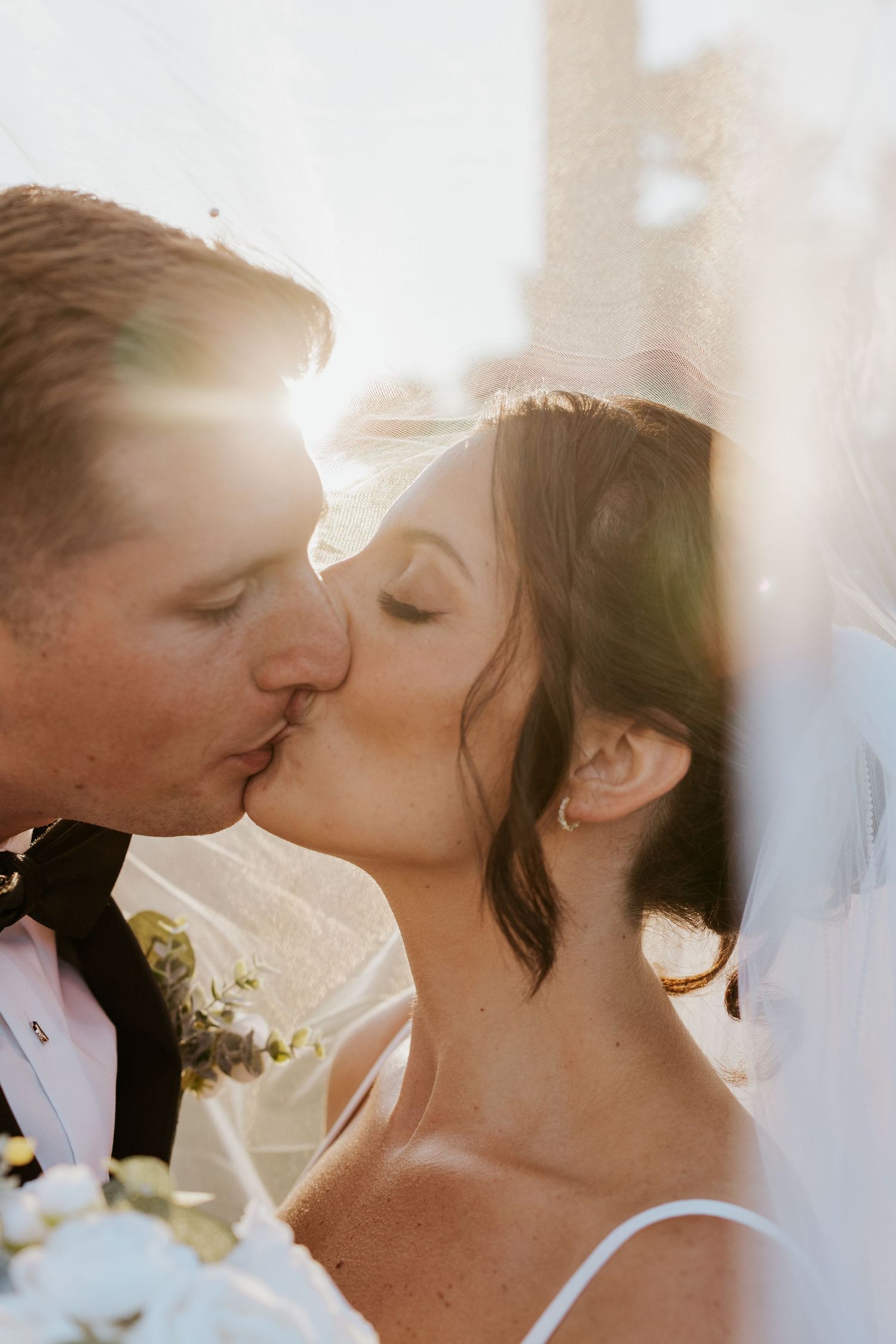 Bride and groom share a romantic kiss under veil in golden sunlight.