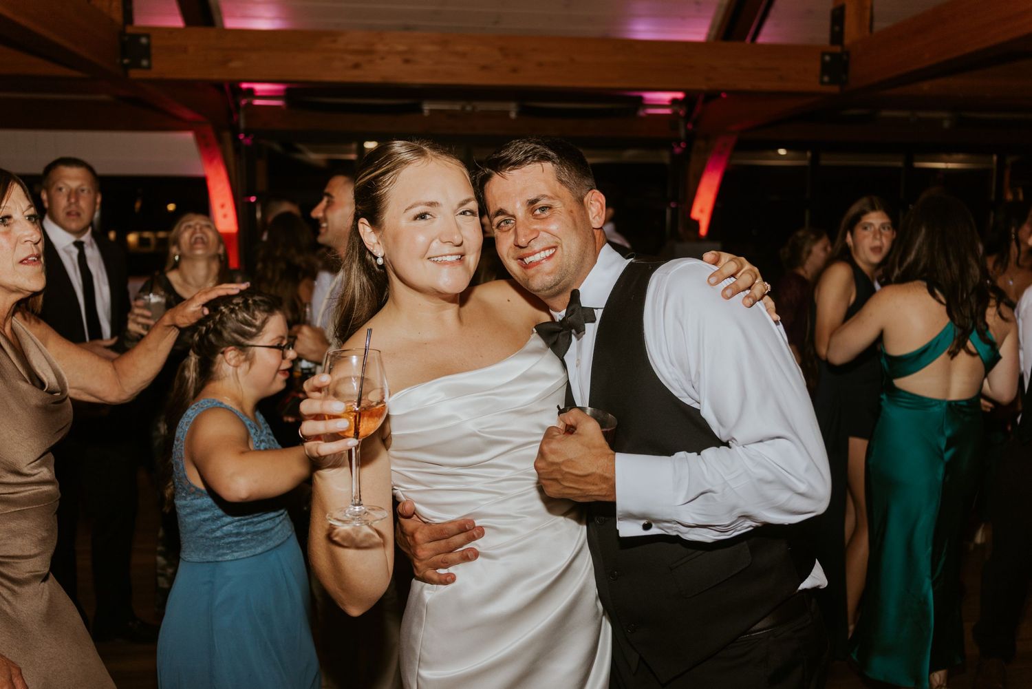 Bride holding champagne and groom smiling together on the dance floor surrounded by wedding guests.
