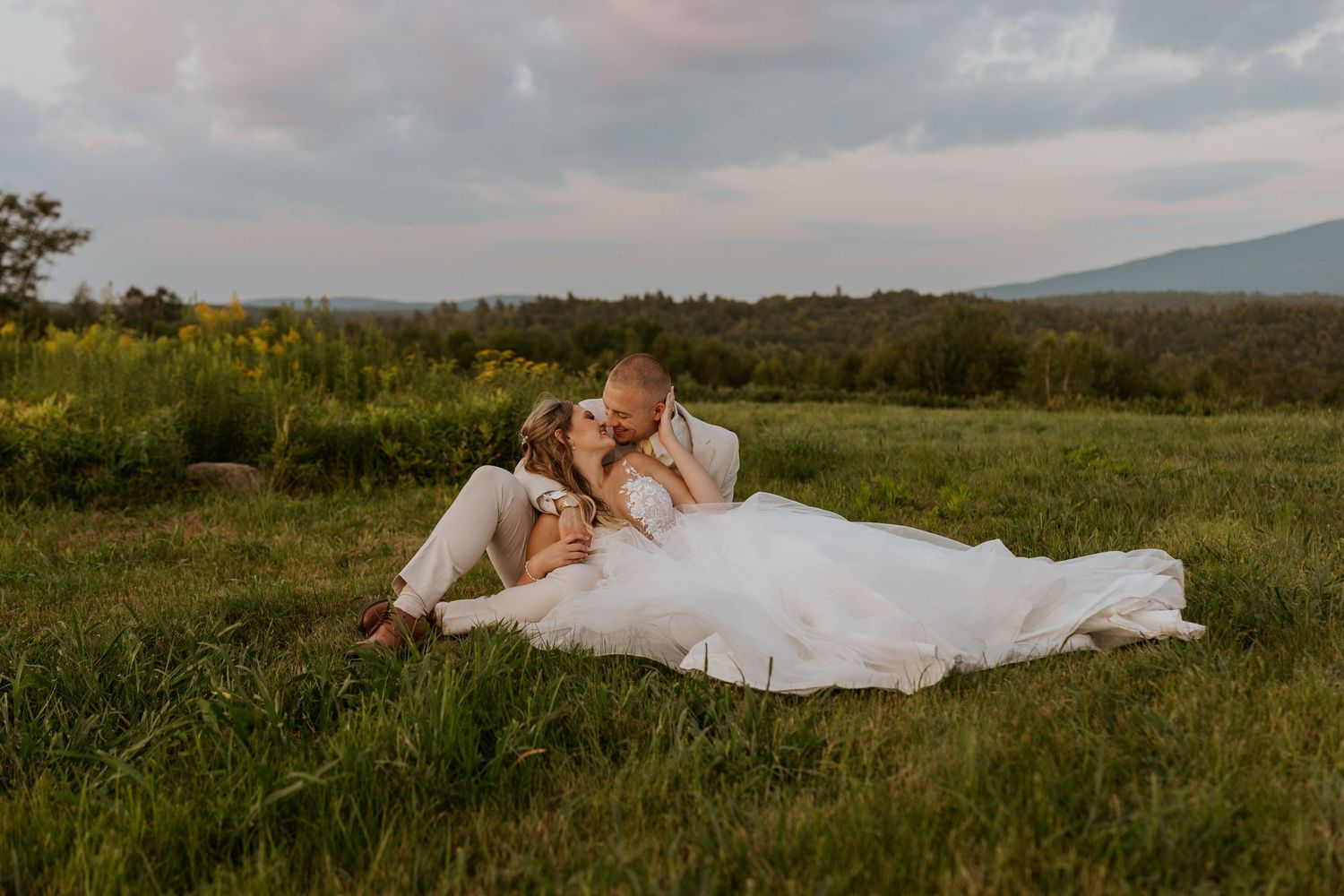 Bride and groom sit together in lush green field kissing with scenic mountain landscape at golden hour.