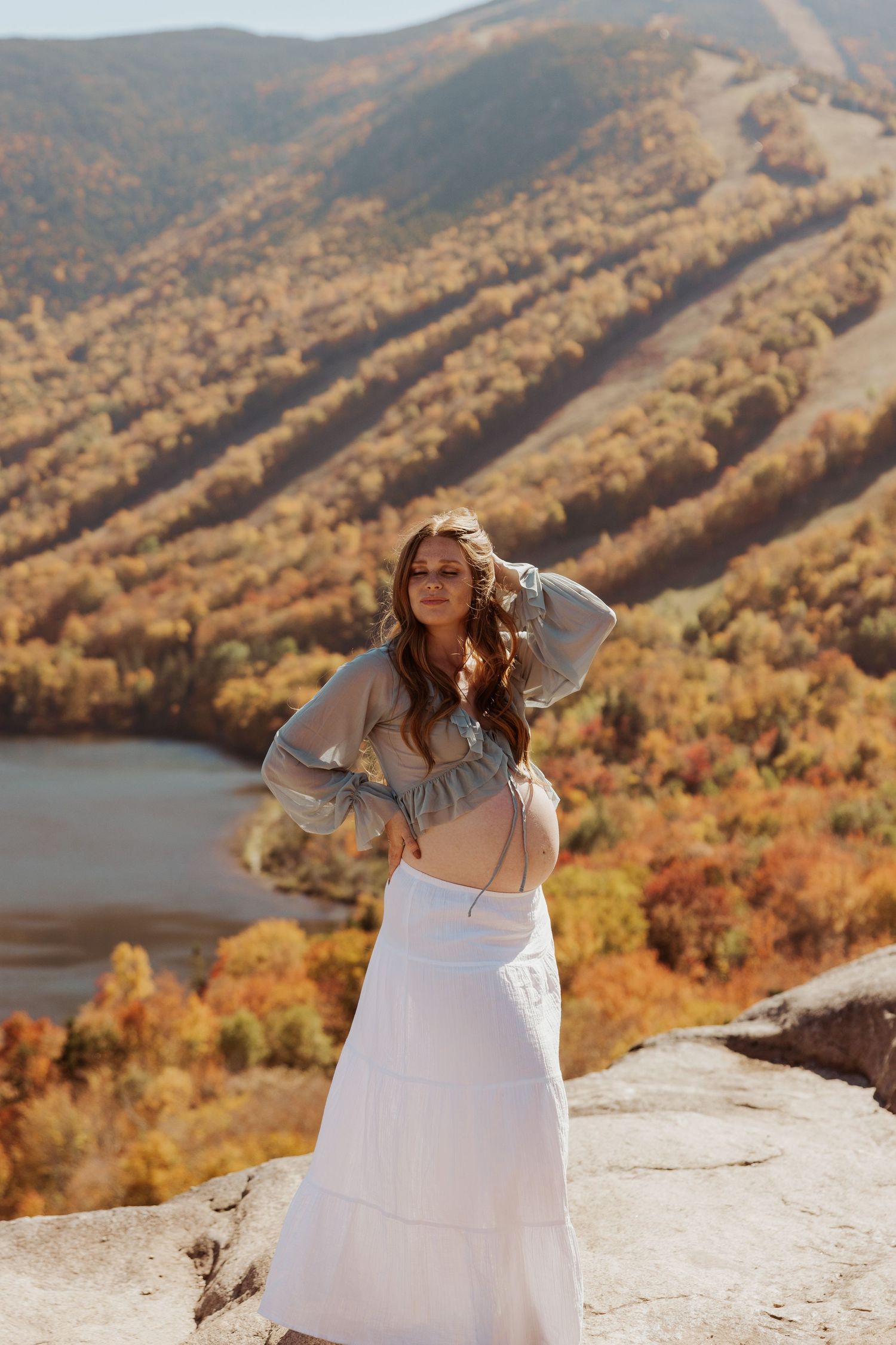 Pregnant woman in sheer top and white skirt posing on mountain overlook with autumn foliage view.