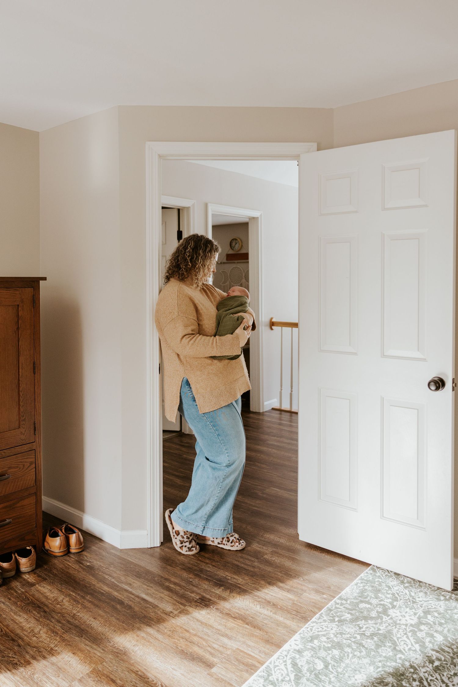 Mother carrying newborn baby in green swaddle walking through doorway of cozy home interior.