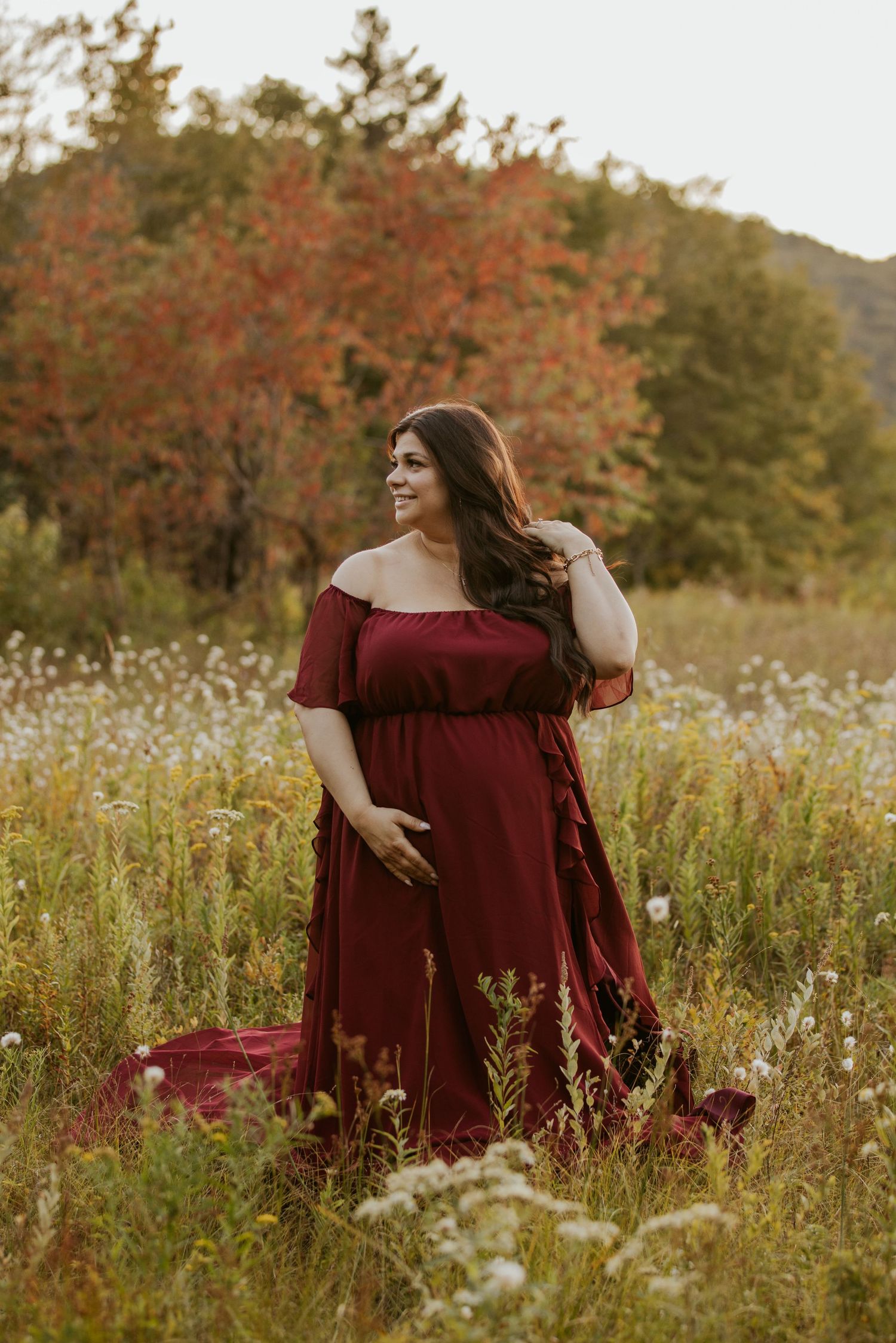 Maternity photo of woman in red gown posing in meadow surrounded by colorful fall foliage.