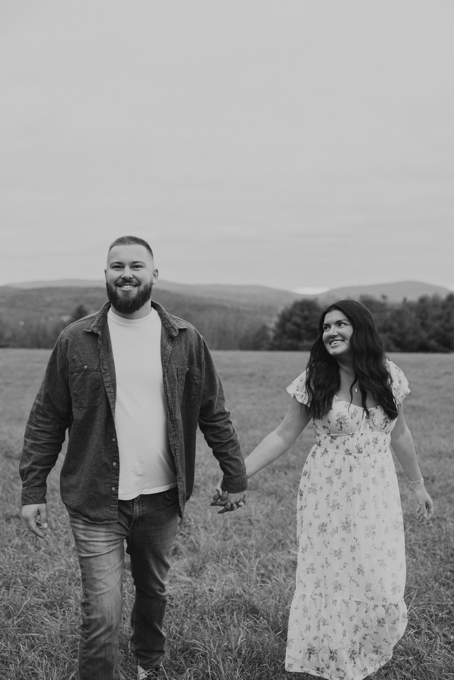 Black and white photo of laughing couple holding hands while walking in open meadow.