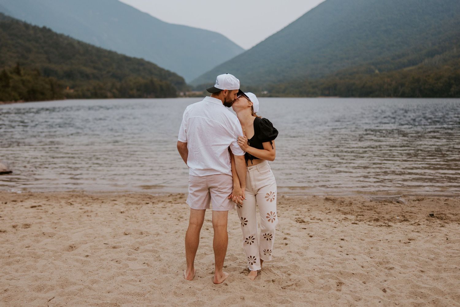 Couple embracing from behind on a sandy beach with a serene mountain lake backdrop.